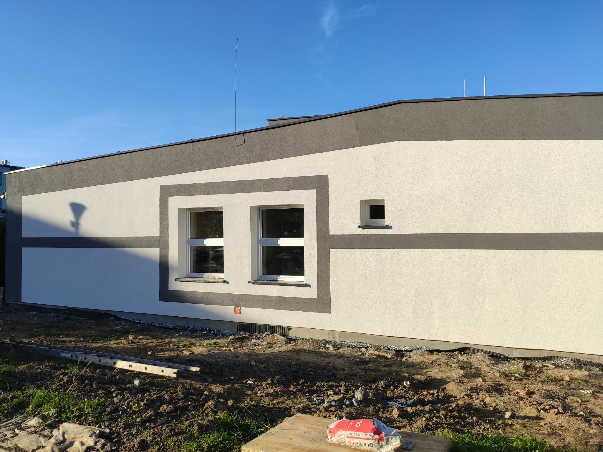Świeżo pomalowana elewacja budynku jednorodzinnego z szarymi akcentami, widoczne dwa okna i małe okienko wentylacyjne, teren wokół jeszcze niezagospodarowany, leży drabina i worek zaprawy.