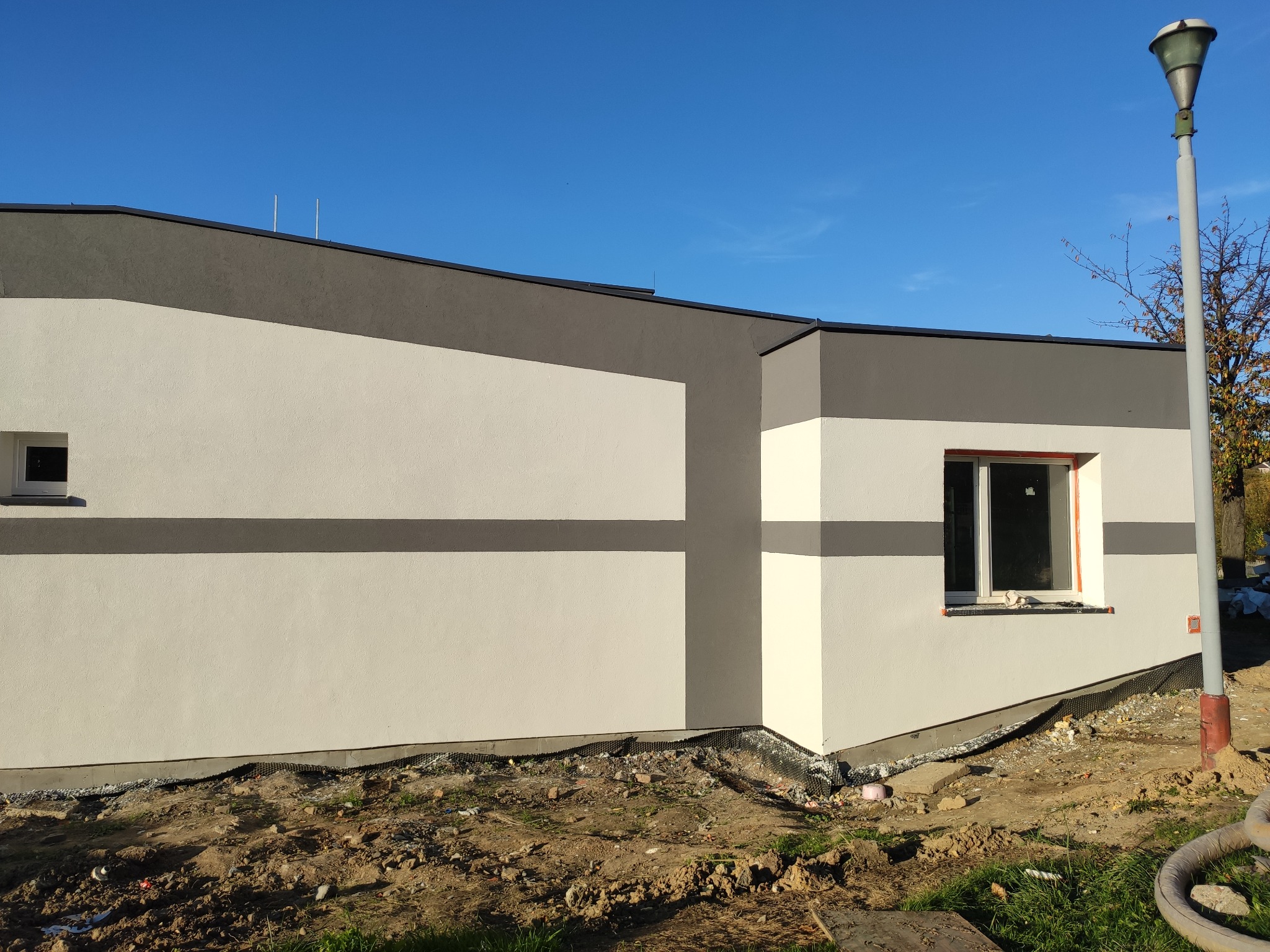 Nowoczesny dom jednorodzinny z elewacją w odcieniach szarości i bieli, widoczne okno z pomarańczową ramą, teren wokół domu w trakcie prac wykończeniowych, lampa uliczna w tle.