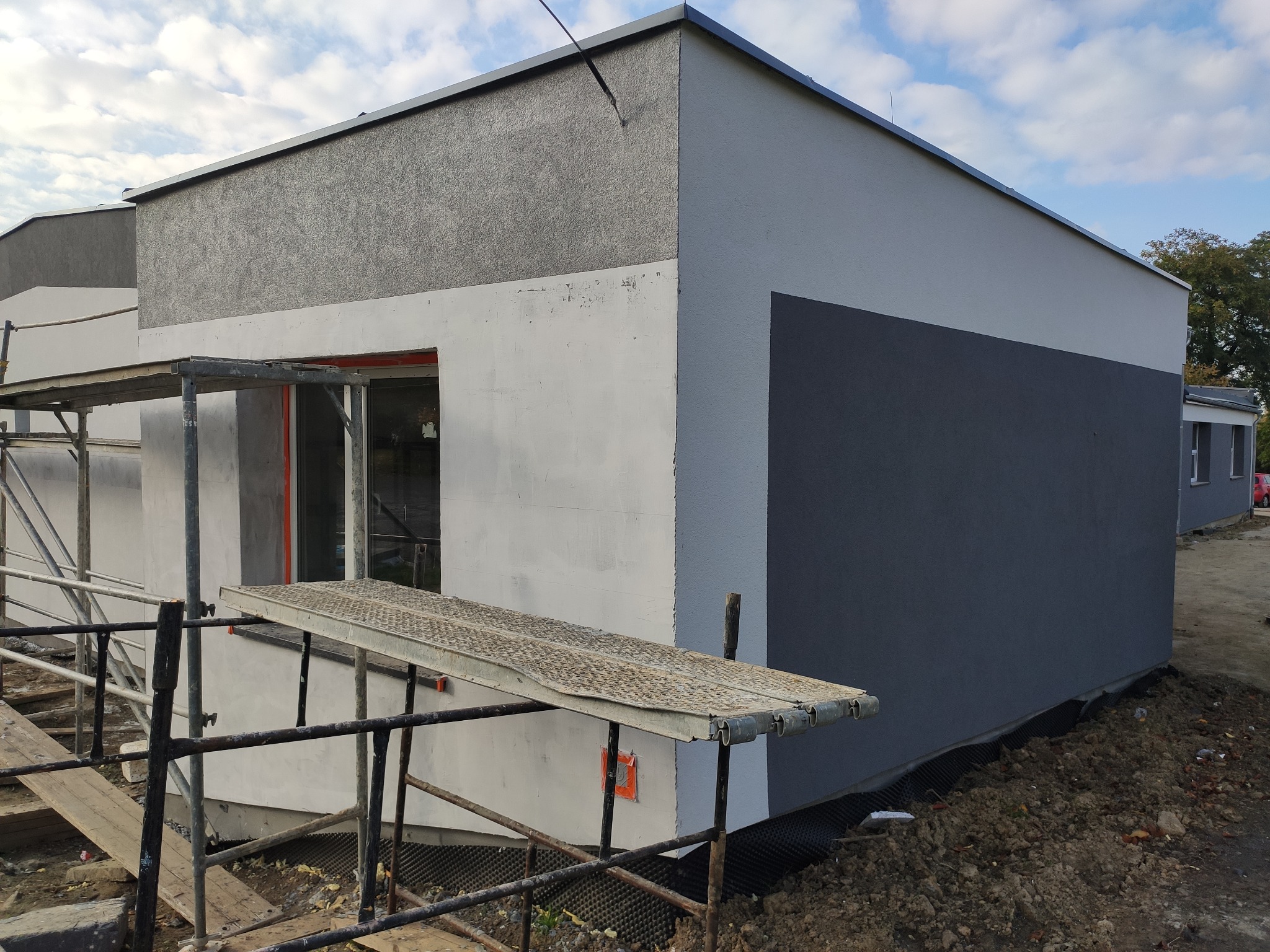 Budynek w trakcie prac elewacyjnych: ocieplony fundament, częściowo pomalowana ściana, rusztowanie wzdłuż budynku. Widoczne nierówności na surowej elewacji i fragmenty niezabezpieczonej folii...