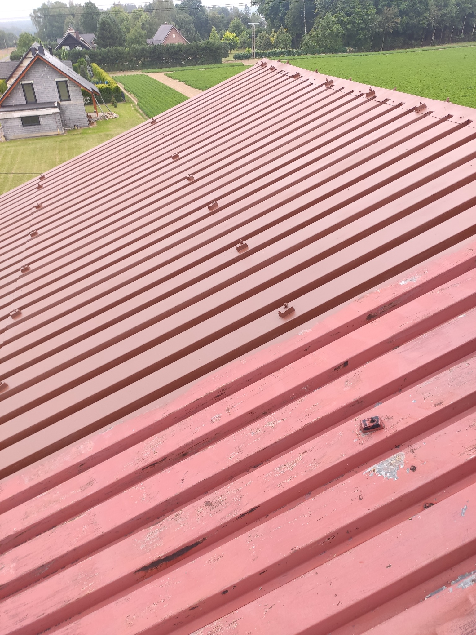 Czerwony blaszany dach z widocznymi mocowaniami, częściowo zniszczoną farbą i widokiem na zielone pola oraz dom w tle.
