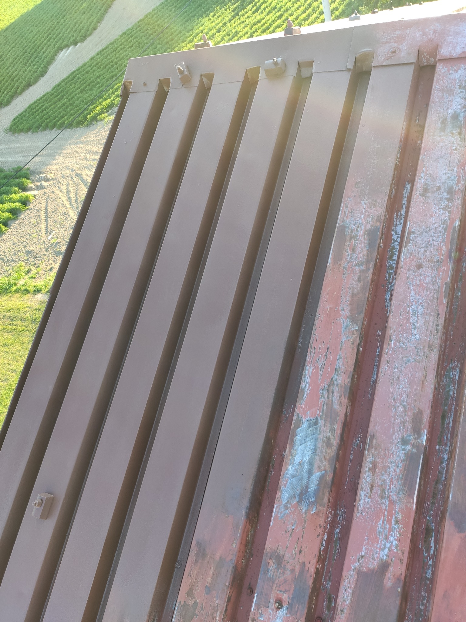 Metalowa konstrukcja dachu z widocznymi śladami korozji i łuszczącej się farby w prawym górnym rogu, na tle zielonego pola uprawnego i błękitnego nieba.