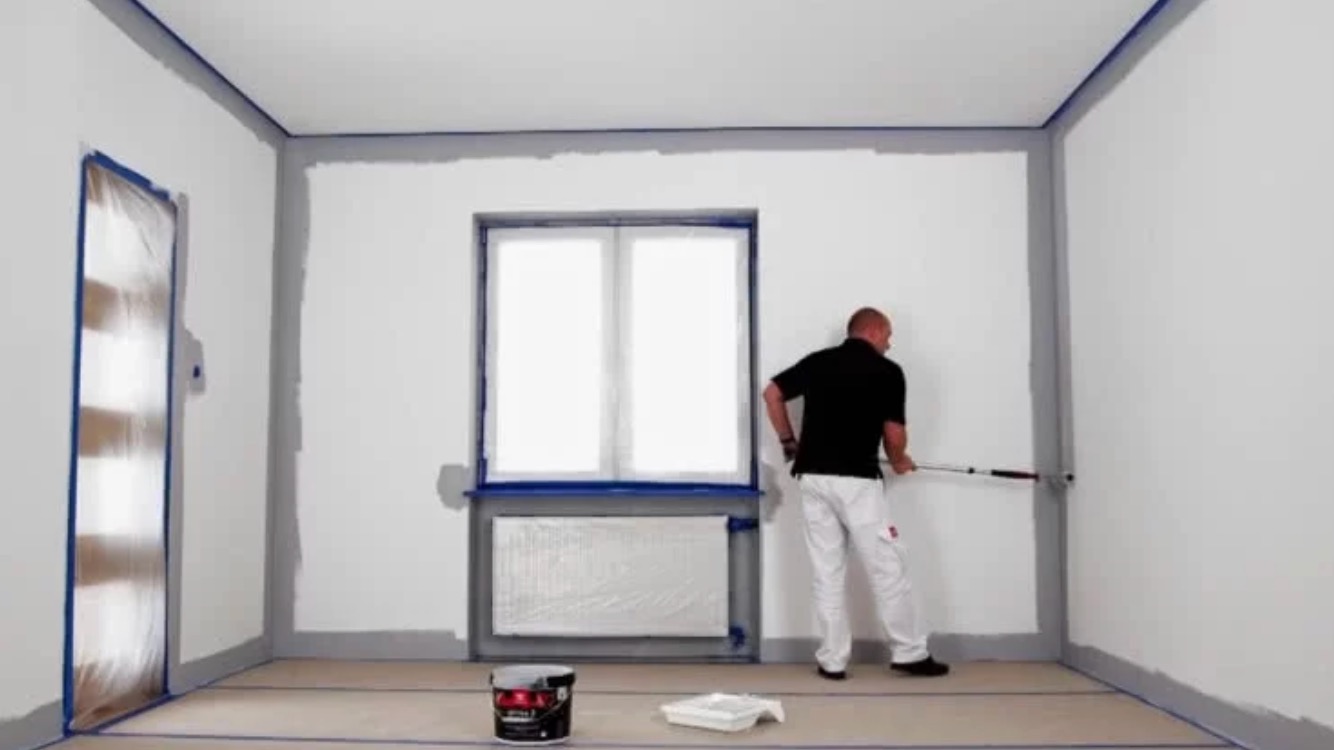 Mężczyzna maluje ścianę wałkiem teleskopowym na szaro, zabezpieczone okno i drzwi folią, widoczne wiadro z farbą i kuweta.