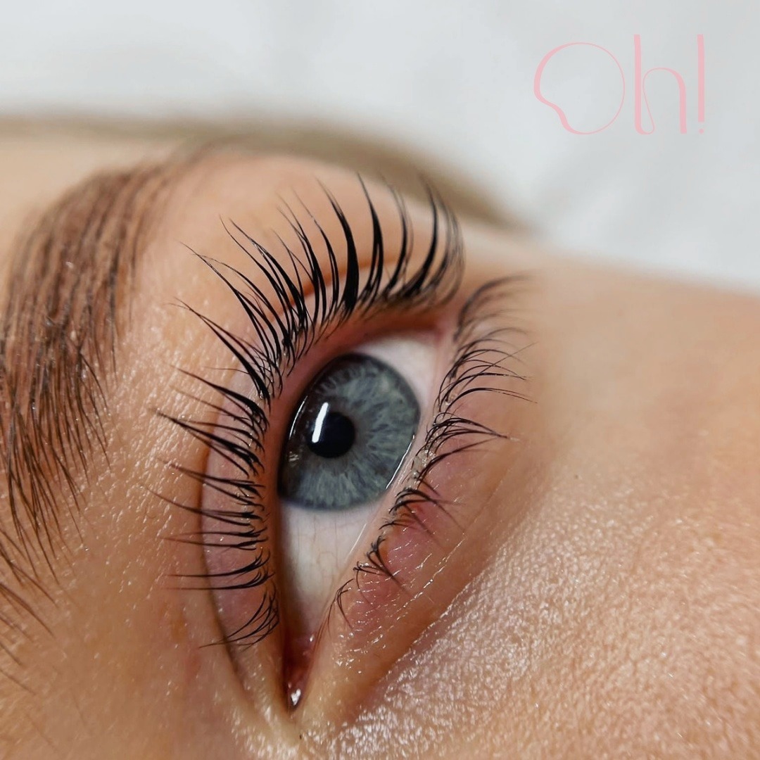 Zbliżenie na oko z niebieską tęczówką i świeżo uniesionymi, czarnymi rzęsami, widoczne delikatne logo w prawym górnym rogu.