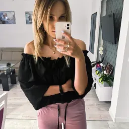 Kobieta robi selfie w lustrze, ubrana w czarną bluzkę z odkrytymi ramionami i różowe spodnie dresowe, widoczna złota biżuteria i manicure w pastelowych kolorach, w tle salon z telewizorem i kwiatami.