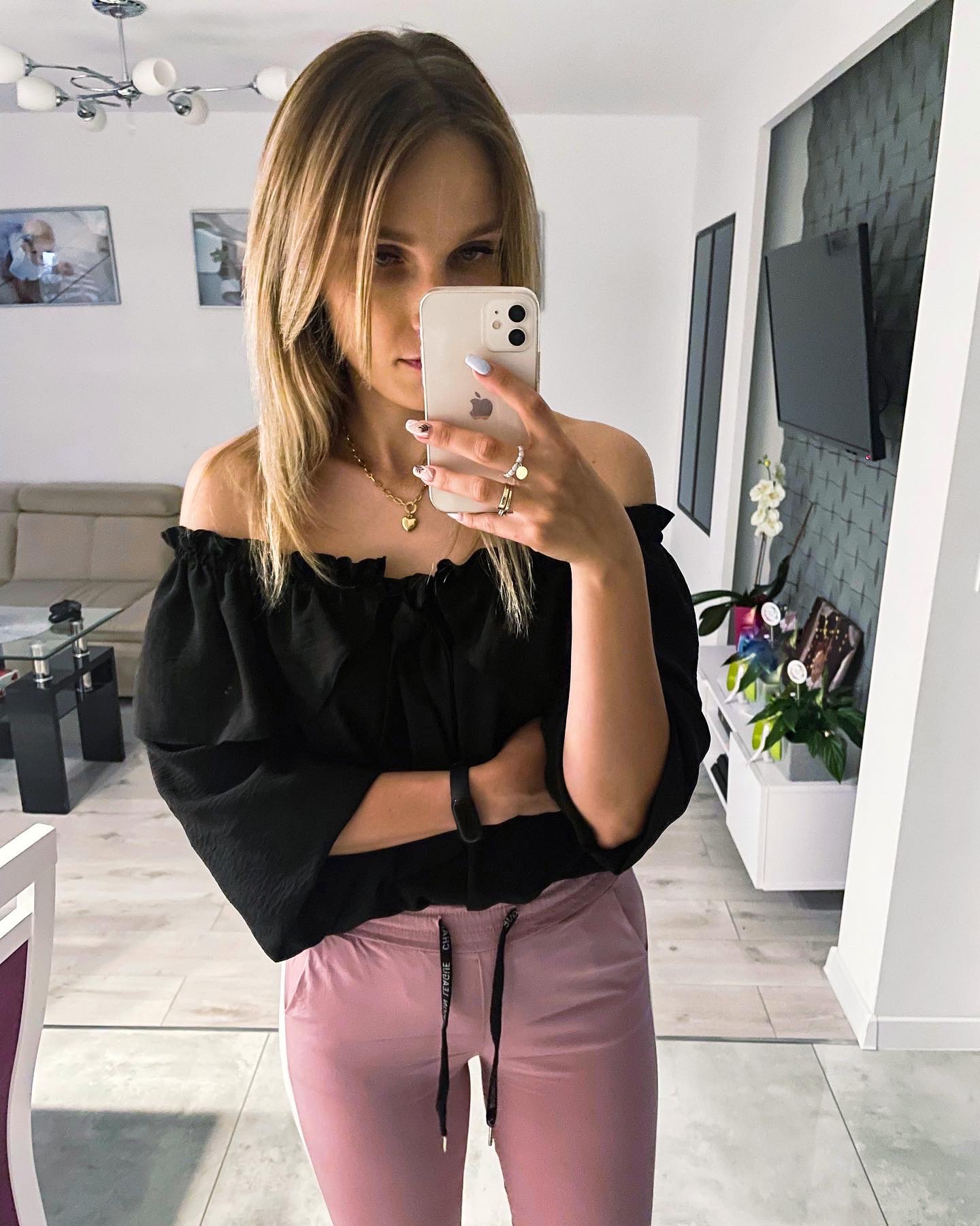 Kobieta robi selfie w lustrze, ubrana w czarną bluzkę z odkrytymi ramionami i różowe spodnie dresowe, widoczna złota biżuteria i manicure w pastelowych kolorach, w tle salon z telewizorem i kwiatami.