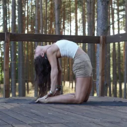 Kobieta wykonuje pozycję odwr&oacute;conego wielbłąda na drewnianym podeście w otoczeniu drzew iglastych, z widocznymi srebrnymi bransoletami na nadgarstkach.