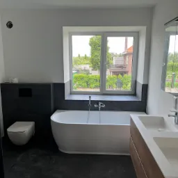 Nowoczesna łazienka z wolnostojącą wanną, podwójną umywalką i oknem z widokiem na zieleń, wykończona w bieli i ciemnej szarości, z elementami drewnopodobnymi.