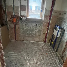 Instalacja ogrzewania podłogowego w trakcie realizacji, widoczne rury grzewcze ułożone na siatce w surowym pomieszczeniu z odsłoniętymi cegłami i instalacją sanitarną.