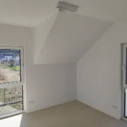 Pusty, jasny pokój z dwoma oknami, skośnym sufitem i nowoczesnym oświetleniem punktowym. Widoczne gniazdka elektryczne na ścianie. Za oknem widok na dom w trakcie budowy.