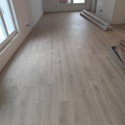 M&E Invest Sp z o.o. usługi elektryczne i glazurnicze - Jasne panele podłogowe ułożone w pokoju z oknami i przygotowanymi ścianami do malowania. Na podłodze leżą zapakowane panele i listwy przypodłogowe.