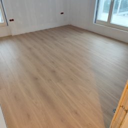 M&E Invest Sp z o.o. usługi elektryczne i glazurnicze - Jasne panele podłogowe ułożone w pustym pokoju z oknami i przygotowanymi ścianami do malowania. Widoczne gniazdka elektryczne i fragment futryny.