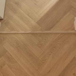 M&E Invest Sp z o.o. usługi elektryczne i glazurnicze - Parkiet ułożony w jodełkę francuską, z widoczną listwą przypodłogową. Jasne drewno, naturalne usłojenie. Ujęcie z góry, fragment wykończonego wnętrza.