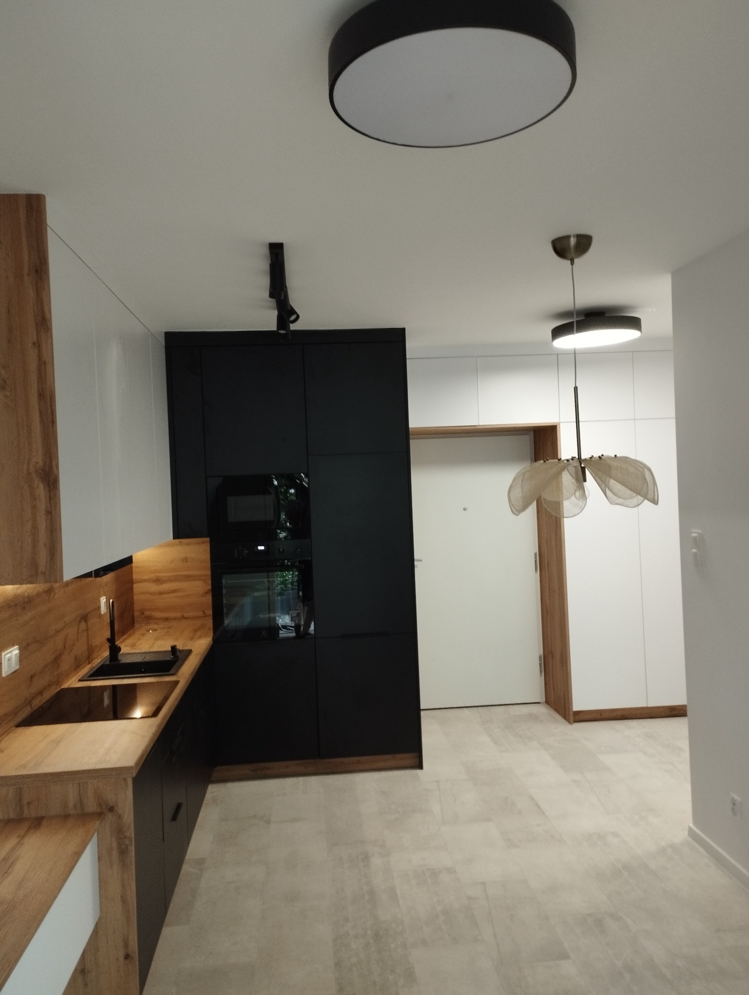Nowoczesna kuchnia z czarnymi szafkami, drewnianym blatem i białymi ścianami, oświetlona trzema lampami sufitowymi, widok na przedpokój z białymi szafami i drewnianymi akcentami.