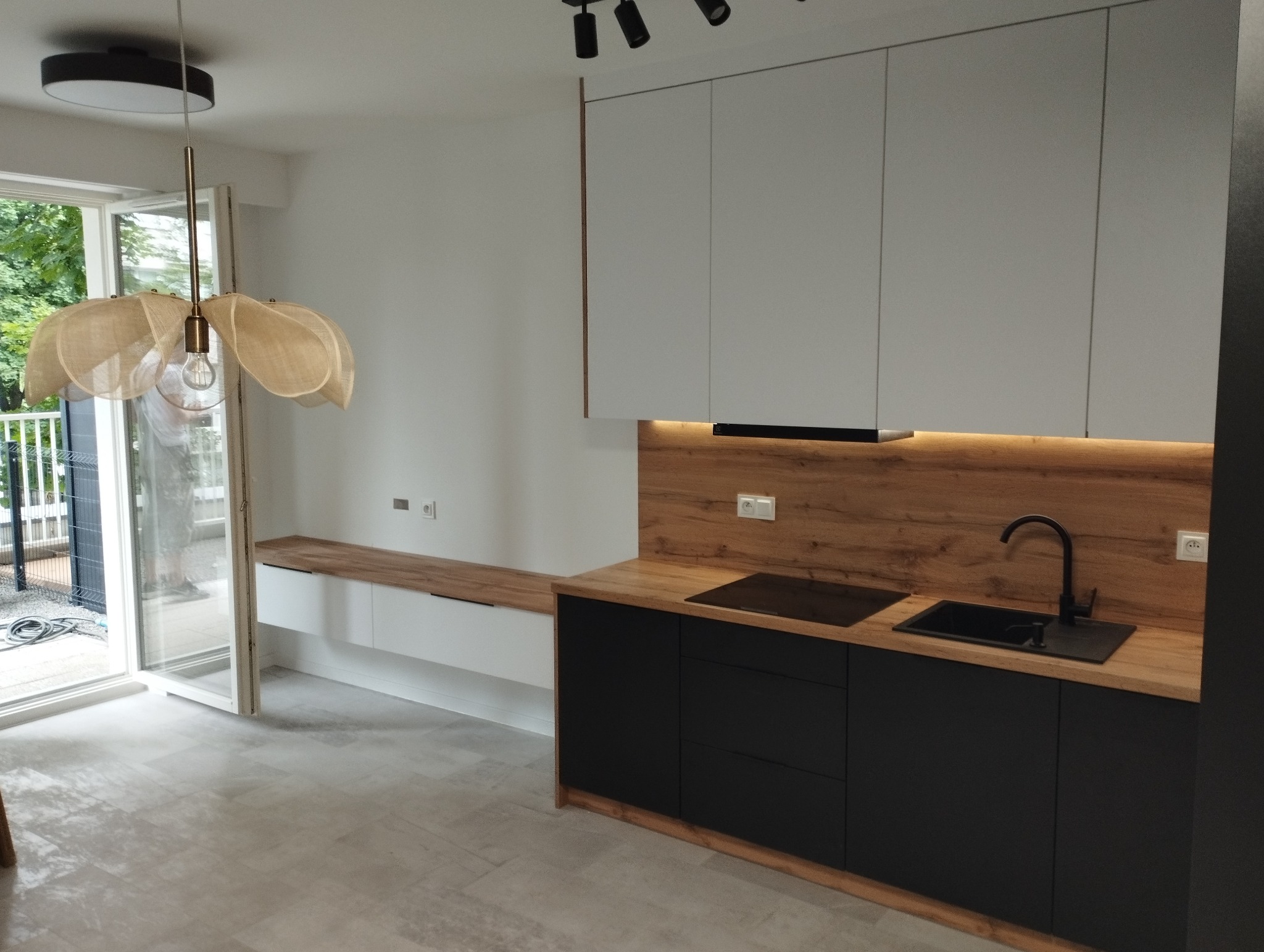Nowoczesna kuchnia z czarnymi szafkami, drewnianym blatem i panelem ściennym, oświetleniem LED pod szafkami wiszącymi, widoczna lampa wisząca z abażurem z naturalnego materiału i wyjście balkonowe.