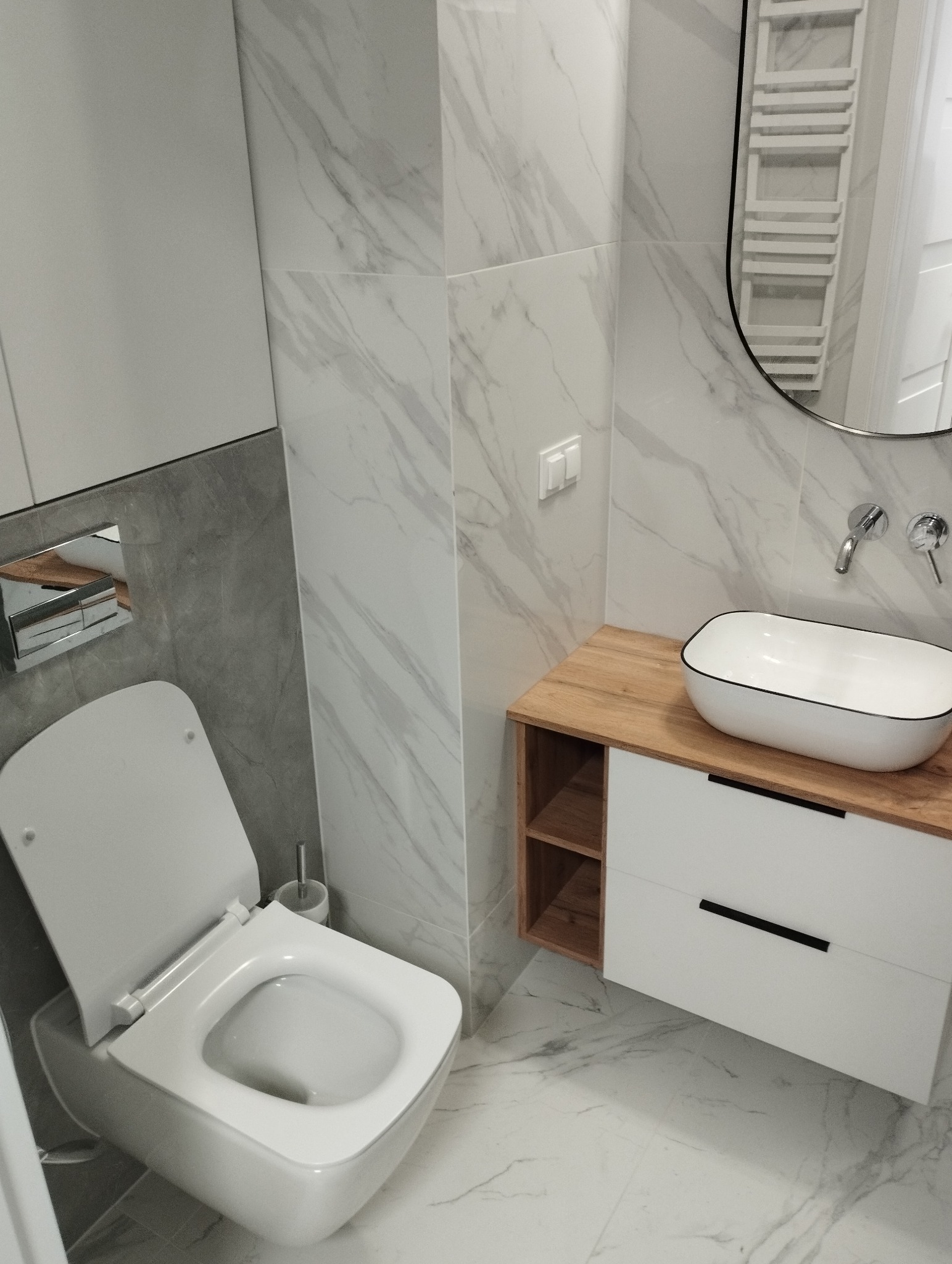 Nowoczesna łazienka z wiszącą miską WC, umywalką nablatową na drewnianym blacie i marmurowymi płytkami na ścianach.