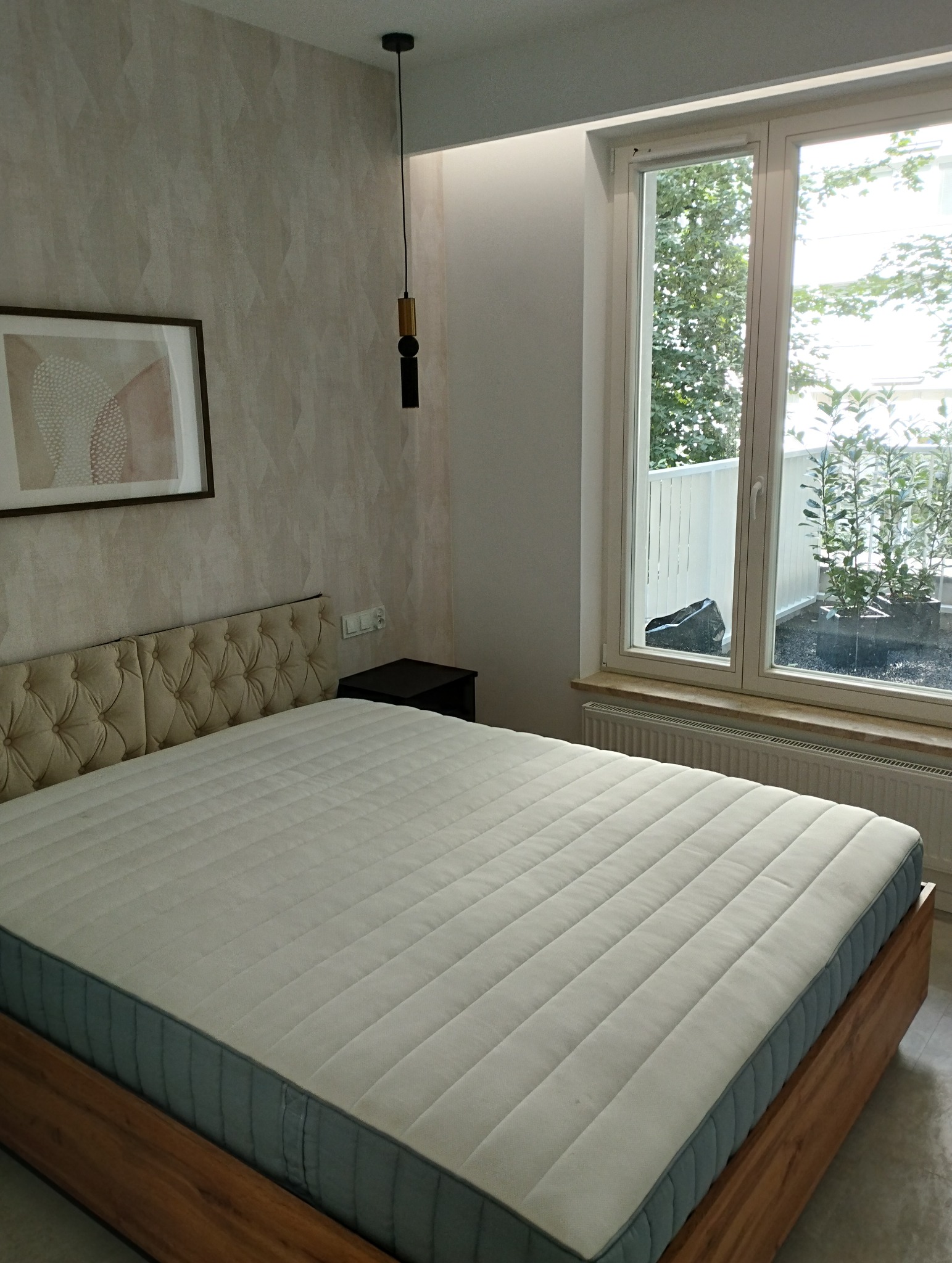 Nowoczesna sypialnia z łóżkiem z pikowanym zagłówkiem, drewnianą ramą i widokiem na balkon z roślinami przez duże okno.