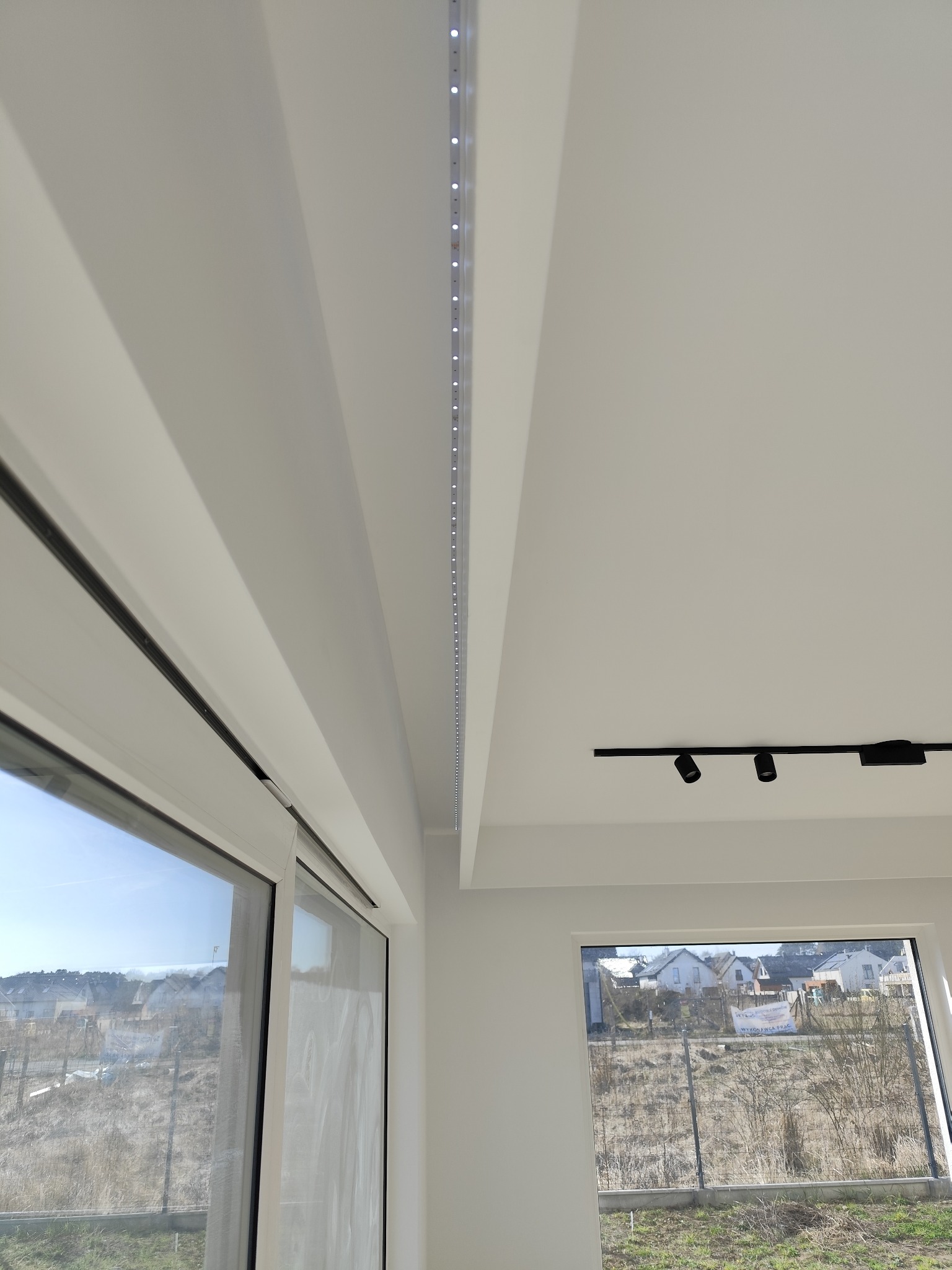 Minimalistyczne wnętrze z białymi ścianami, oświetleniem LED w listwie sufitowej i reflektorami na szynie, widok na zewnątrz przez okno.