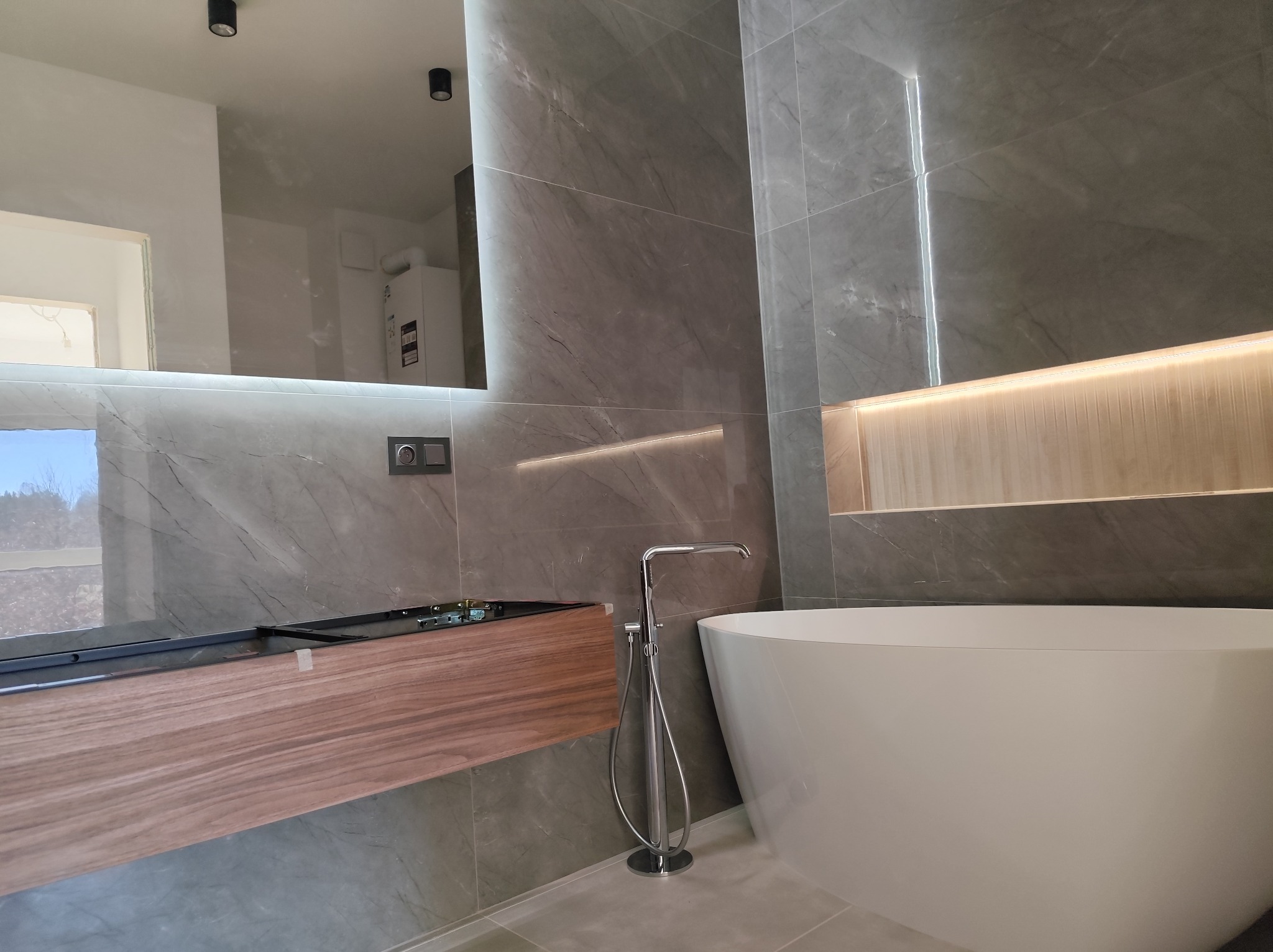 Nowoczesna łazienka z wanną wolnostojącą, szafką pod umywalką z drewnianym fornirem i szarymi płytkami imitującymi marmur, oświetlenie LED wzdłuż ściany i pod lustrem.