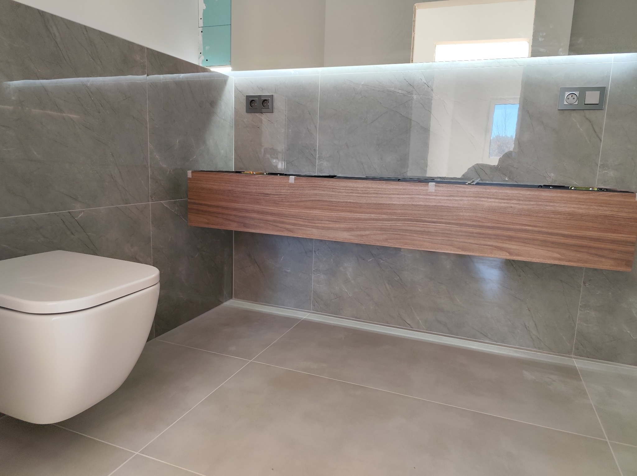 Nowoczesna łazienka z szarymi marmurkowymi płytkami, podwieszaną toaletą i drewnianą szafką pod umywalkę z podświetleniem LED, widoczne gniazdka elektryczne