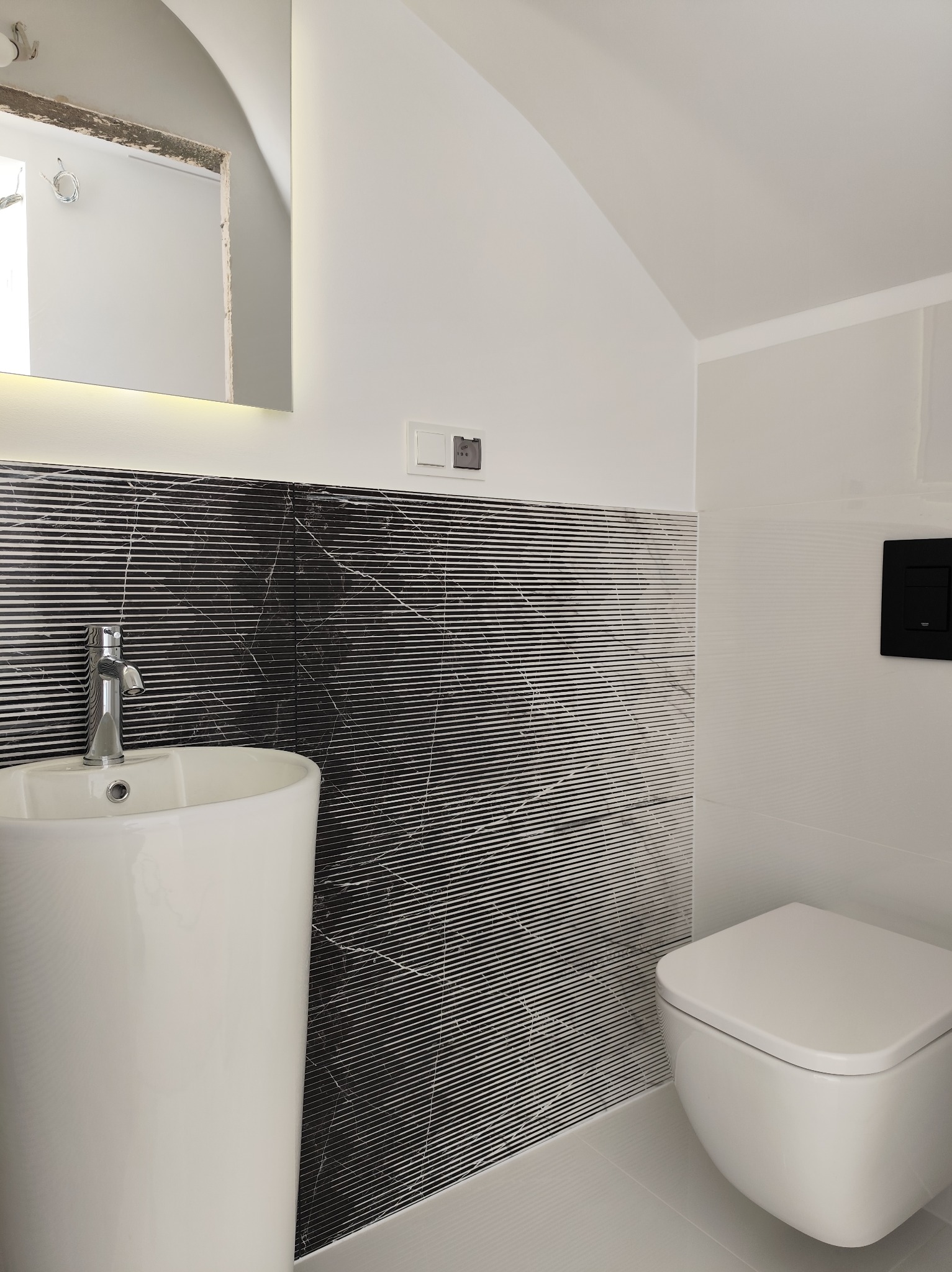 Nowoczesna łazienka z umywalką kolumnową, wiszącą toaletą i czarno-białymi płytkami z poziomymi paskami, oświetlona lustrem LED i z czarnym panelem sterującym spłuczką.