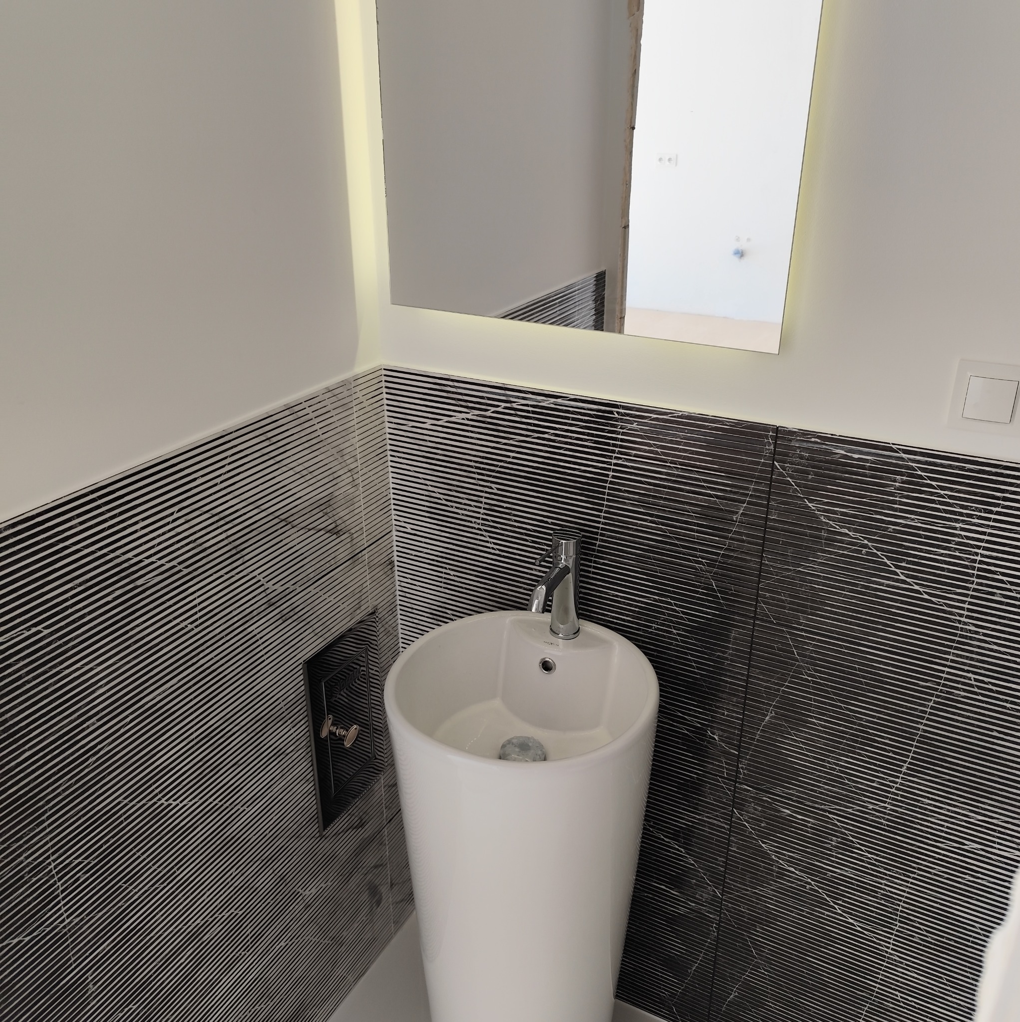 Biała, wolnostojąca umywalka z chromowaną baterią na tle ściany wyłożonej czarno-białymi płytkami w paski, oświetlone lustro z wbudowanym światłem LED.