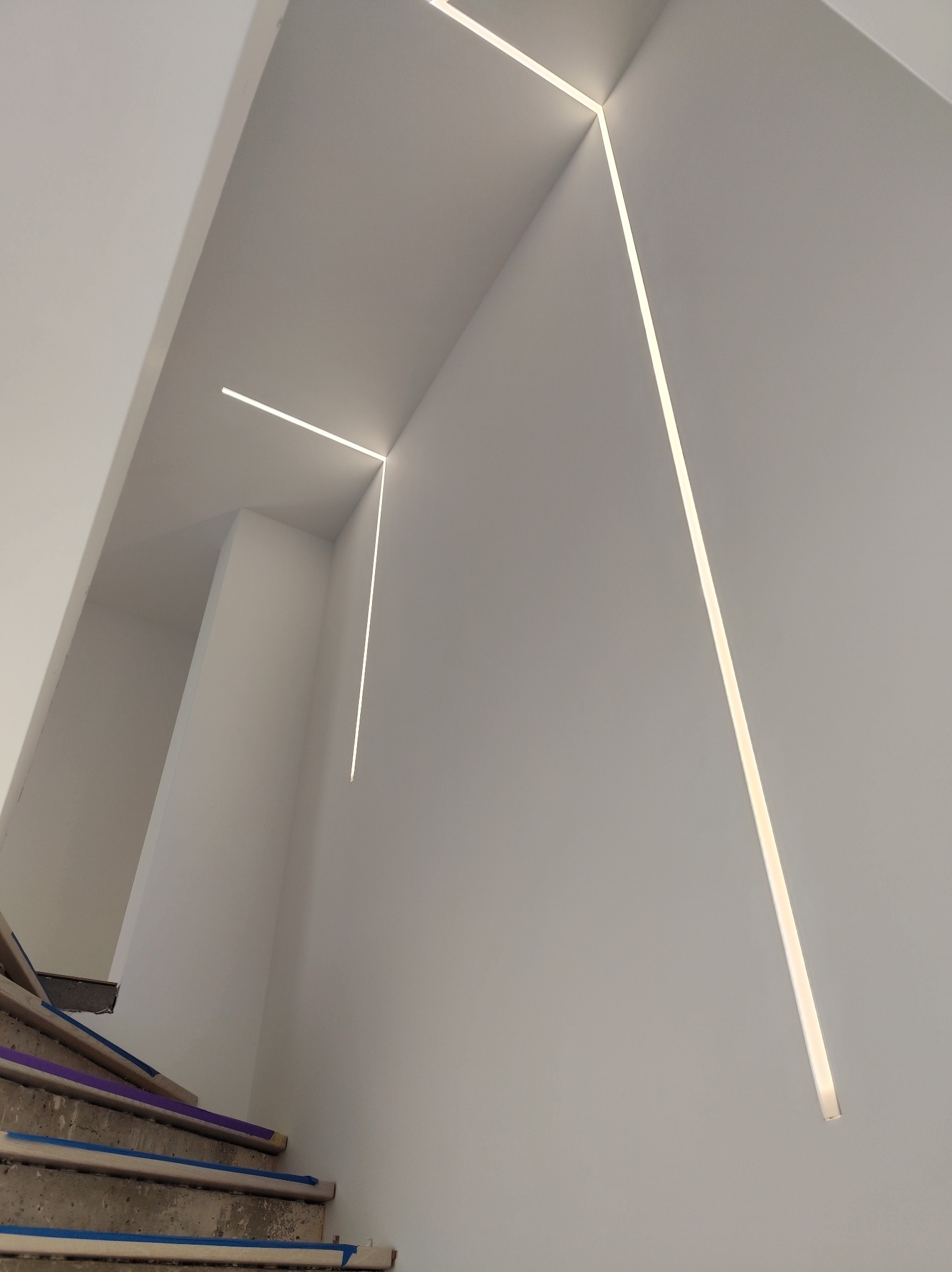 Minimalistyczne oświetlenie schodów z wbudowanymi liniowymi lampami LED w białych ścianach, schody zabezpieczone taśmą malarską.