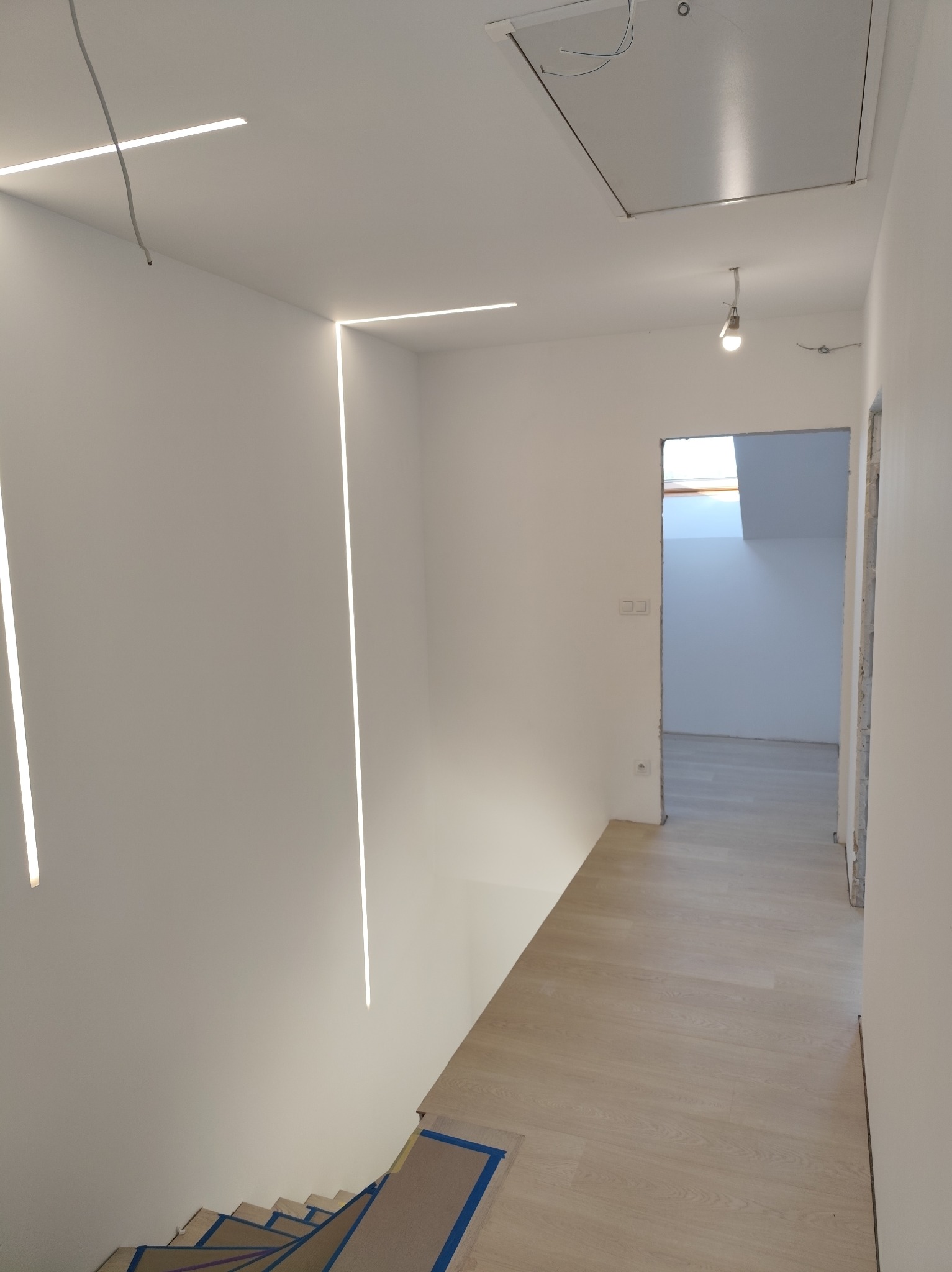 Wnętrze domu w trakcie prac wykończeniowych: białe ściany z wbudowanym oświetleniem LED, zabezpieczone schody z drewnianymi stopniami i fragment widocznej podłogi, sufit z klapą inspekcyjną.