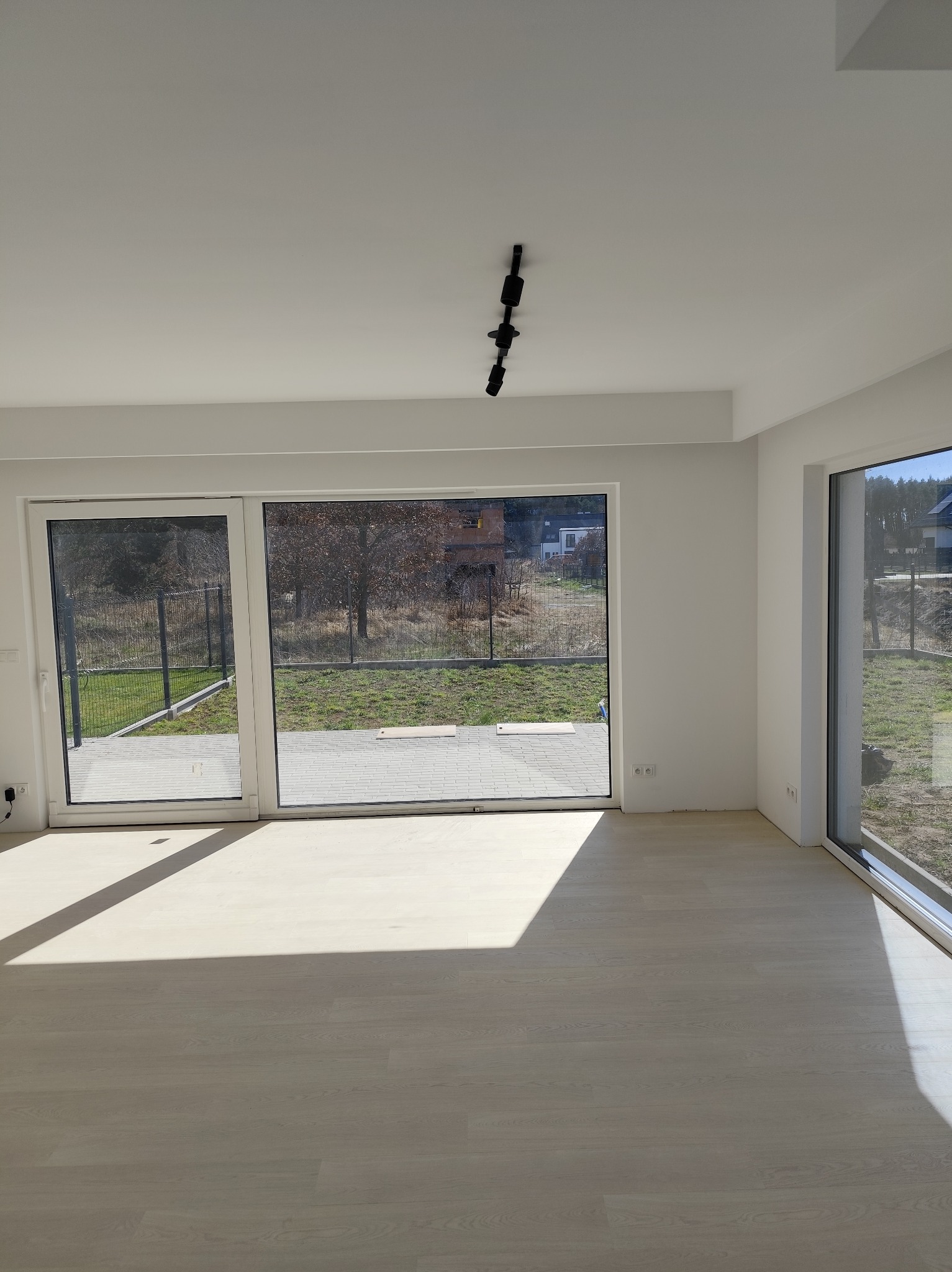 Pusty, jasny pokój z drewnianą podłogą, dużymi przesuwnymi oknami z widokiem na ogród i czarną lampą sufitową.