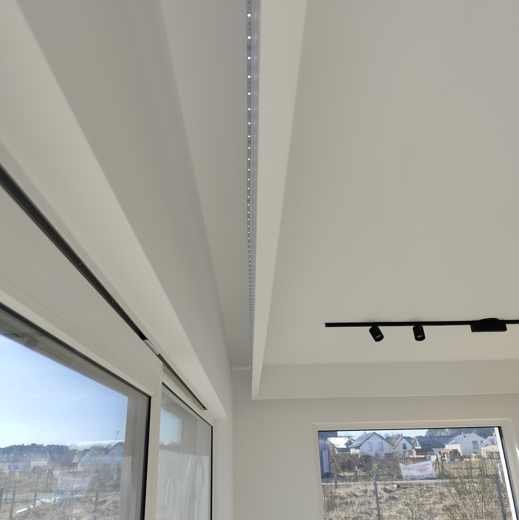 Białe wnętrze z oknem, listwą LED w suficie i czarnymi reflektorami punktowymi na szynie, widok z dołu.