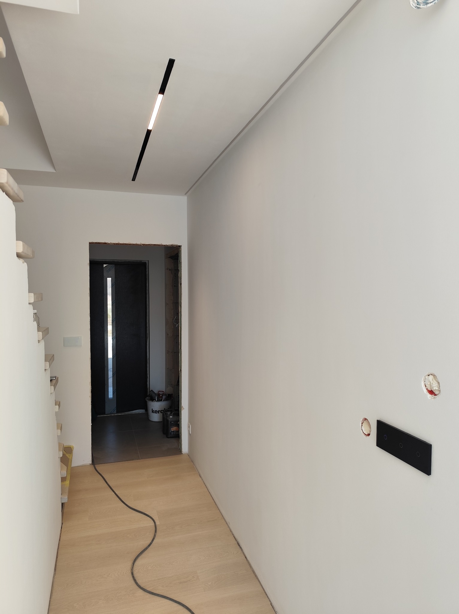 Korytarz w trakcie remontu z nową podłogą, oświetleniem liniowym na suficie i gładzią na ścianach, widoczne puszki elektryczne i przewód na podłodze.