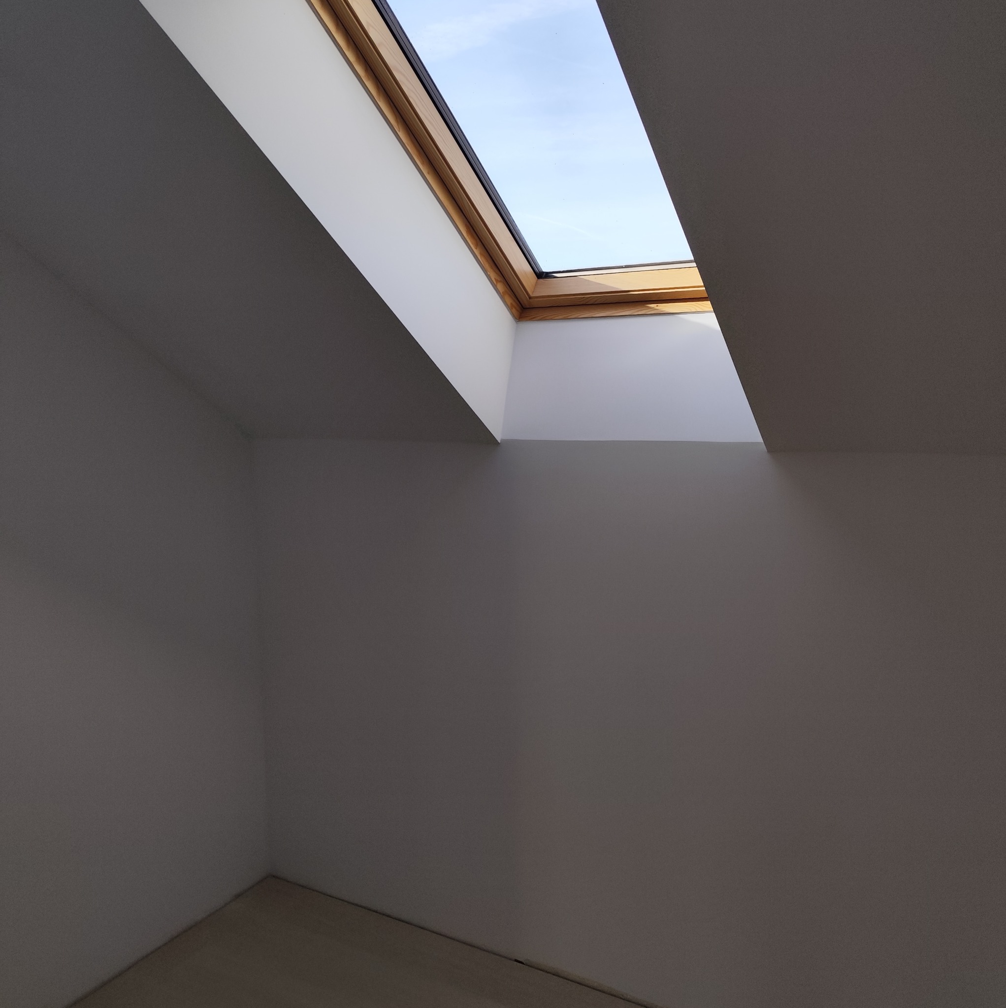 Poddasze ze świeżo pomalowanymi ścianami na biało i szaro, z drewnianą ramą okna dachowego, przez które widać fragment błękitnego nieba.