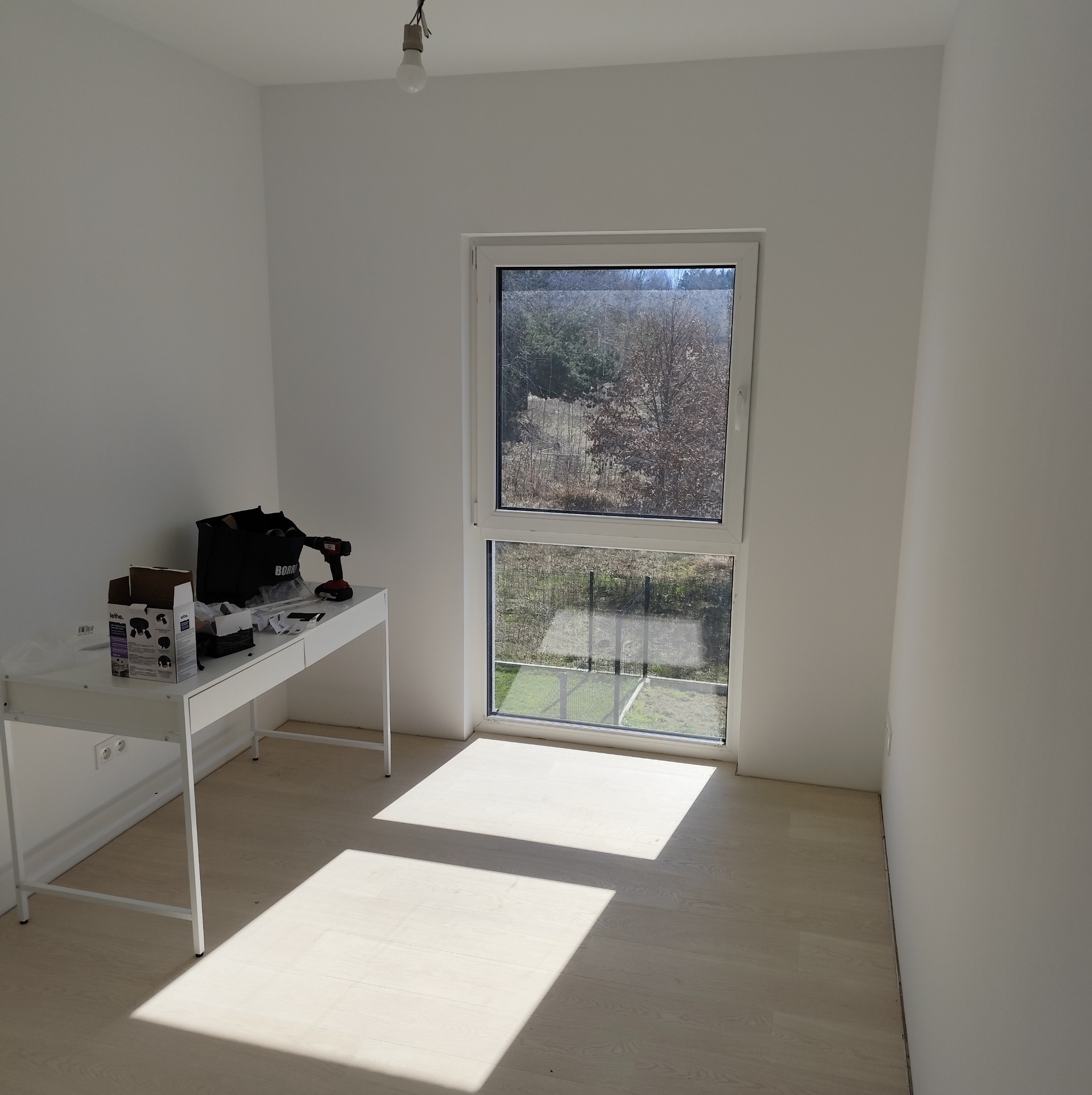 Pusty, jasny pokój z białymi ścianami i oknem z widokiem na drzewa; na biurku narzędzia i akcesoria elektryczne w trakcie pracy.