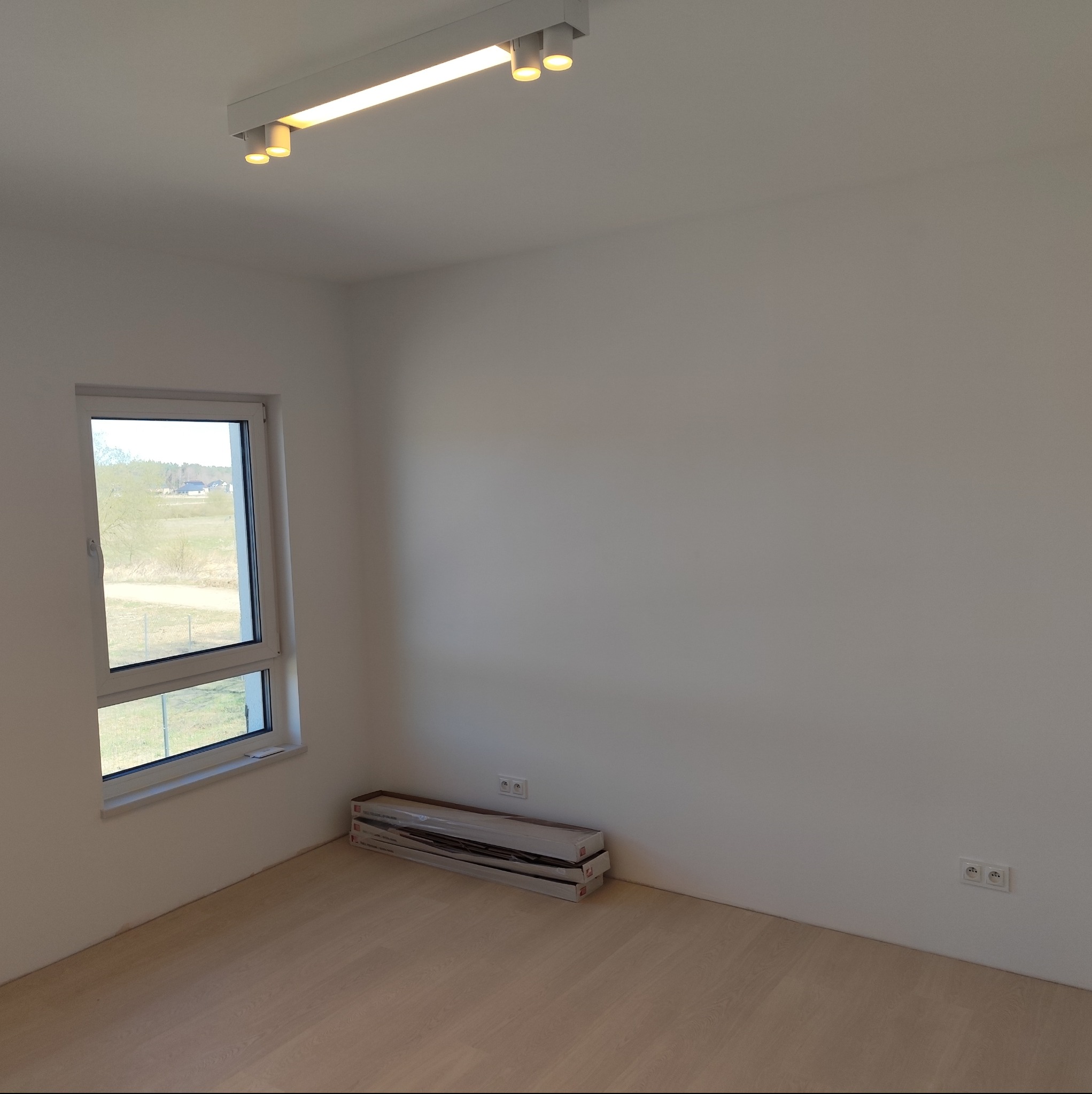 Pusty pokój z jasnymi ścianami i parkietem, okno z widokiem na zewnątrz, na podłodze leżą paczki paneli podłogowych, na suficie biała lampa sufitowa z reflektorami, widoczne gniazdka elektryczne...