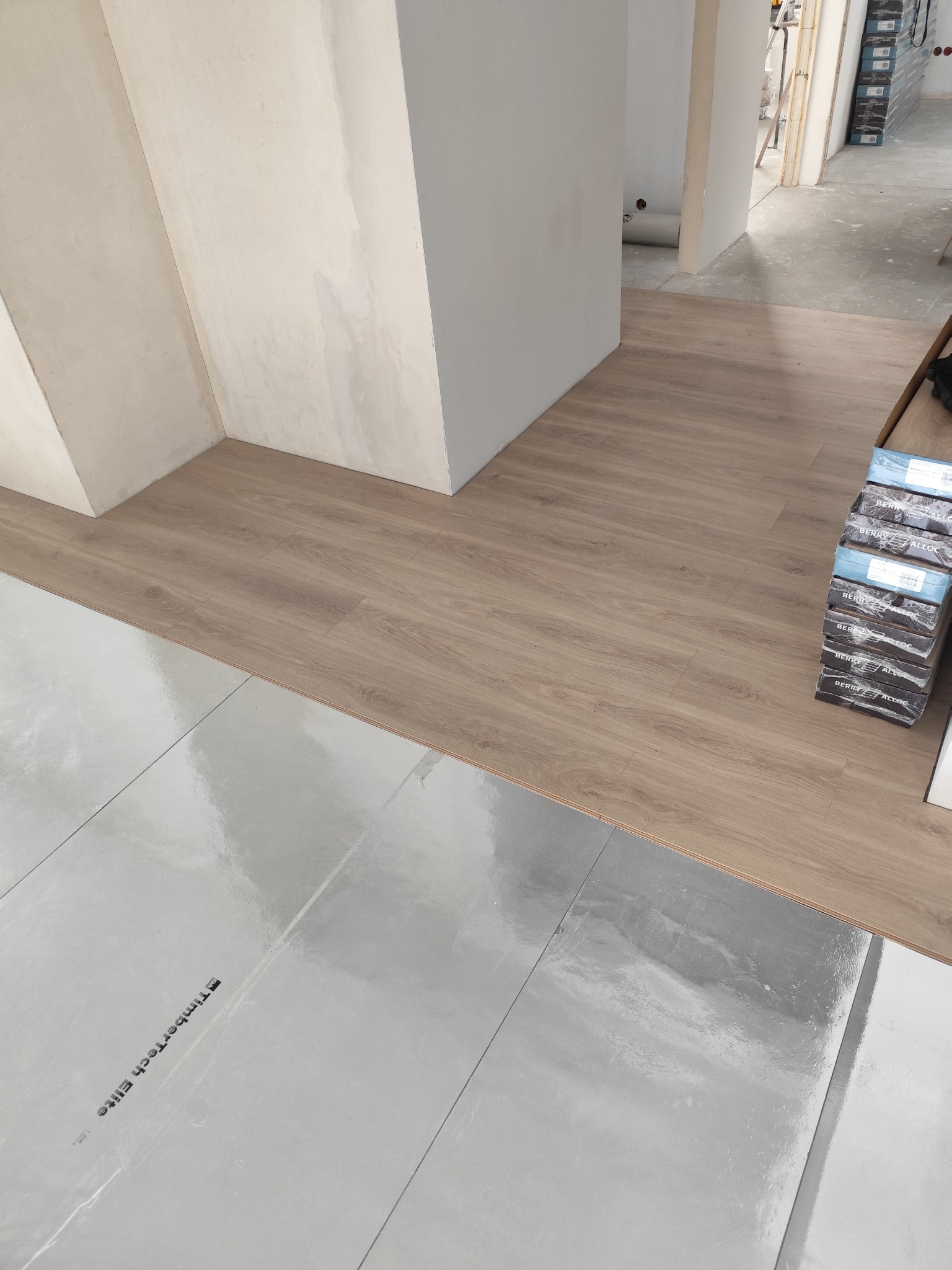 Układanie paneli podłogowych na folii izolacyjnej, widoczne paczki paneli Berry Alloc, fragment ściany w tle. Etap prac wykończeniowych.