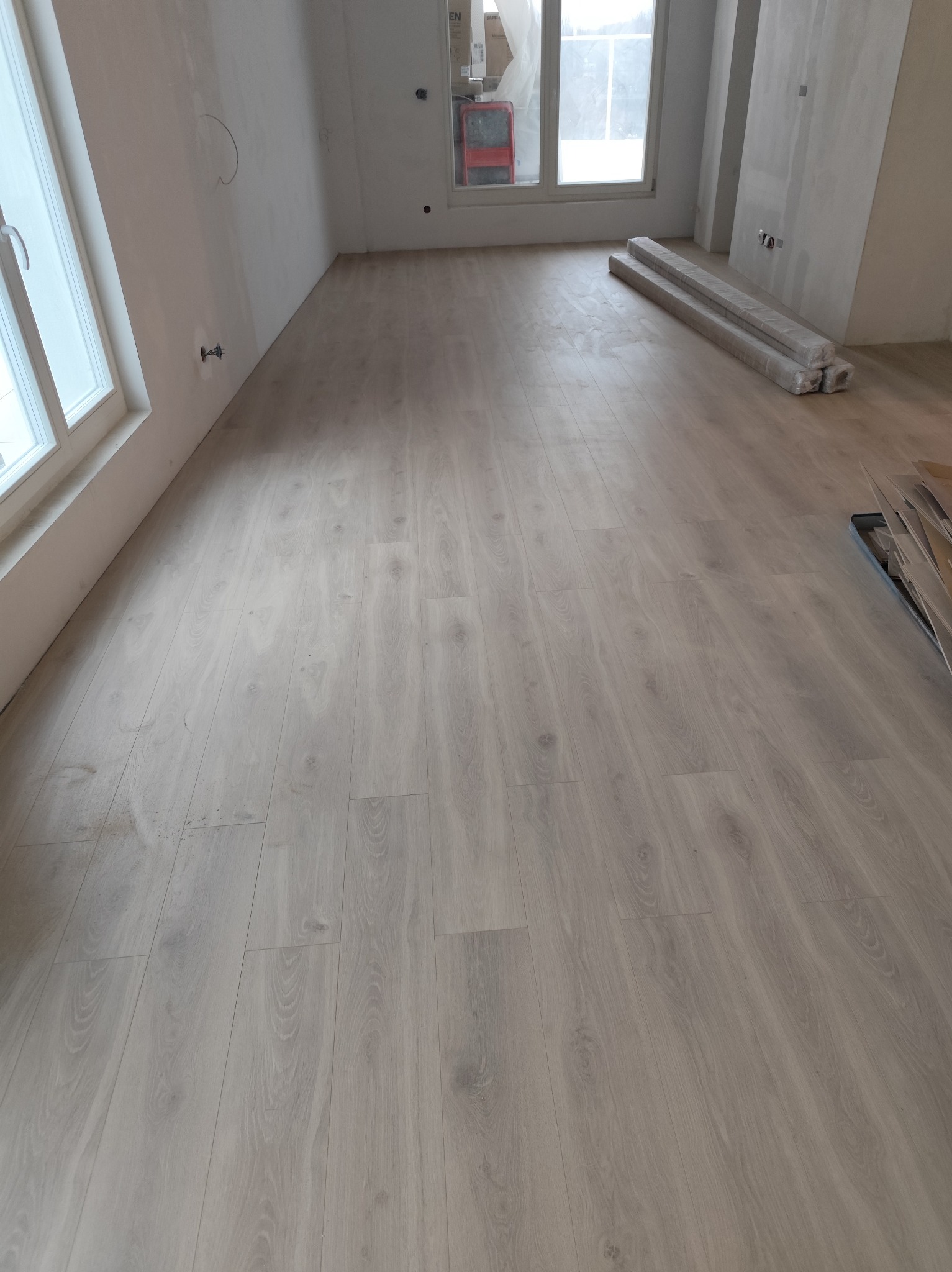 Jasne panele podłogowe ułożone w pokoju z oknami i przygotowanymi ścianami do malowania. Na podłodze leżą zapakowane panele i listwy przypodłogowe.