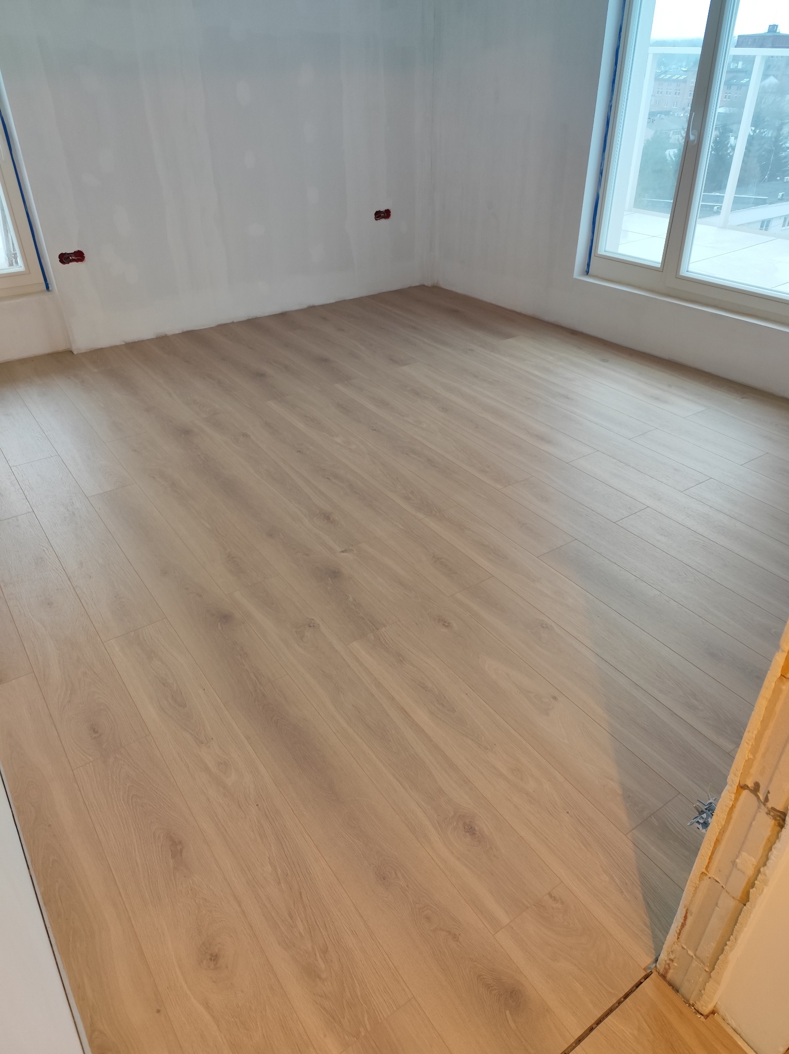 Jasne panele podłogowe ułożone w pustym pokoju z oknami i przygotowanymi ścianami do malowania. Widoczne gniazdka elektryczne i fragment futryny.