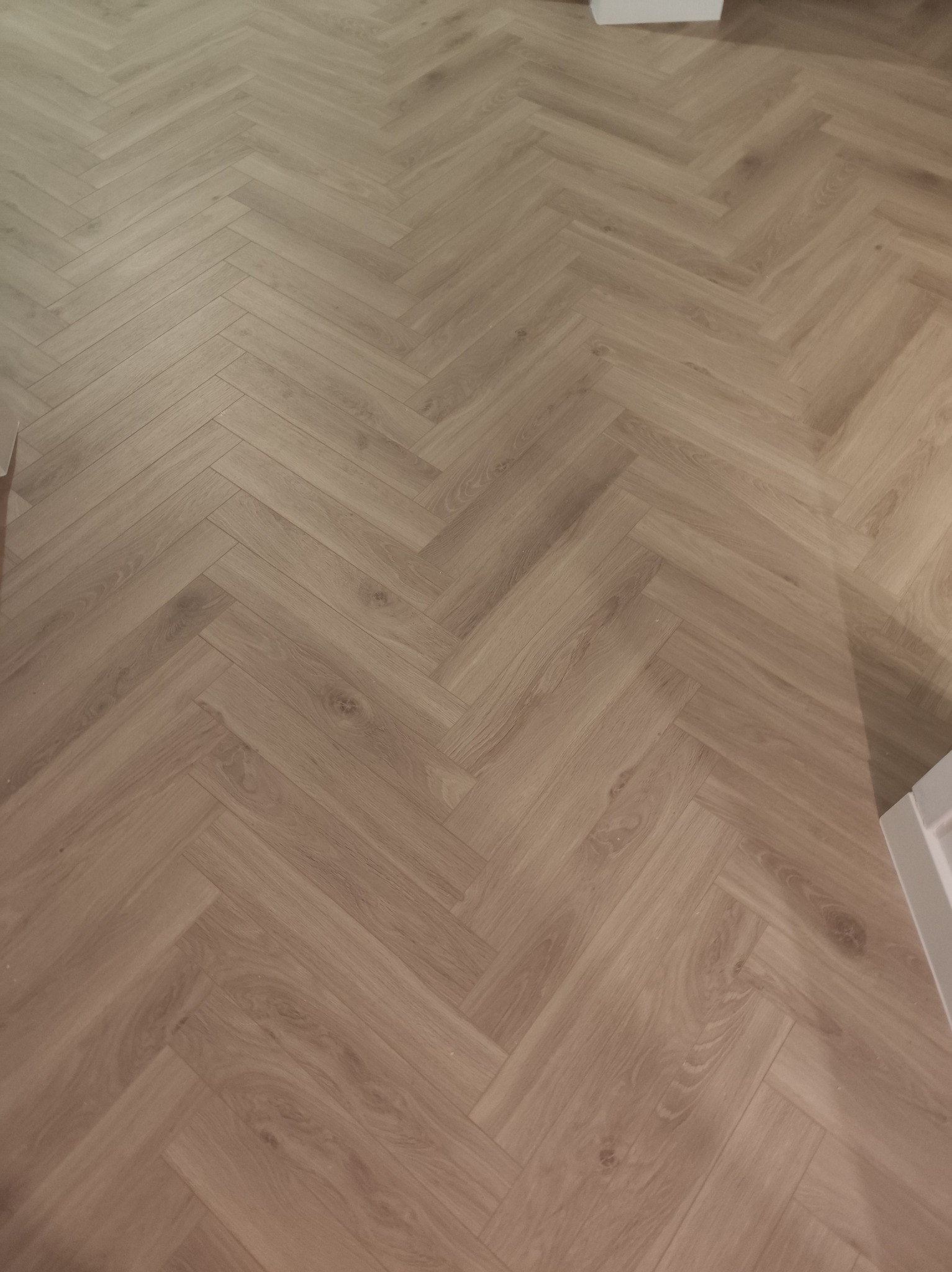 Jasna podłoga ułożona w jodełkę francuską, widok z góry. Parkiet z wyraźnym rysunkiem drewna, delikatne oświetlenie, fragment białej listwy przypodłogowej w prawym dolnym rogu.