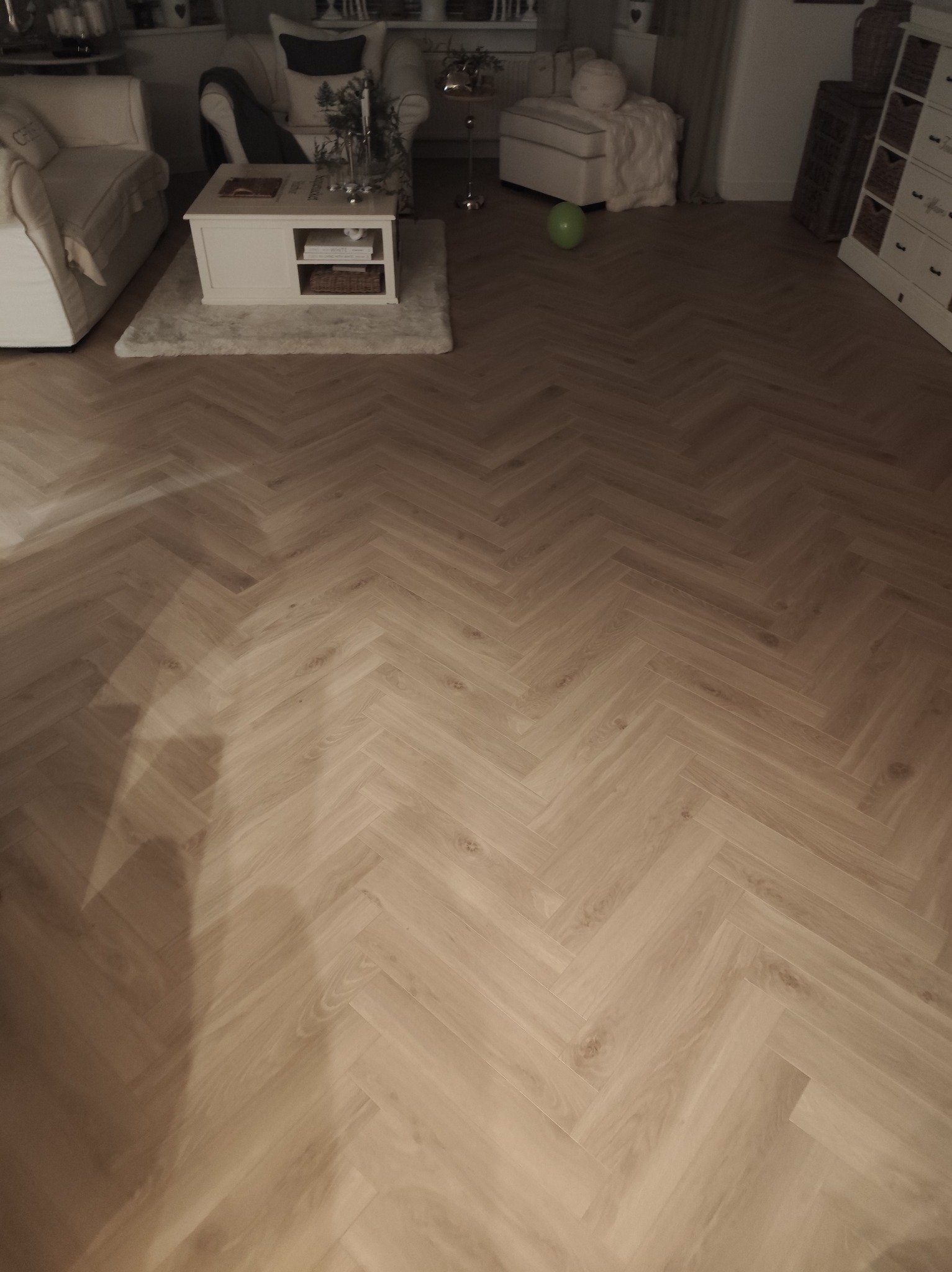 Jasna, drewniana podłoga ułożona w jodełkę francuską, widok z góry. W tle fragment salonu z białymi meblami i zielonym balonem.