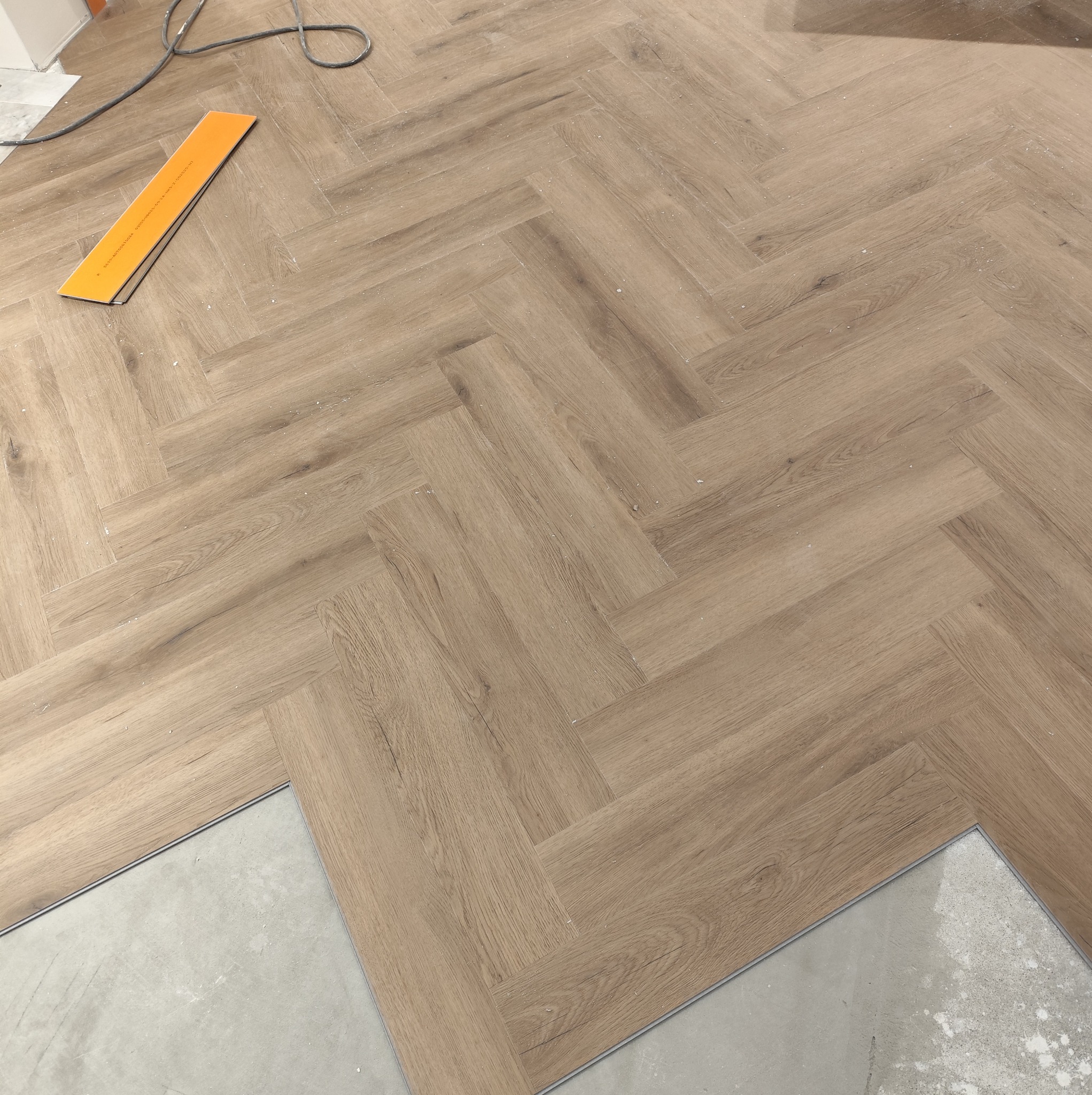 Układanie paneli podłogowych w jodełkę, widoczny fragment surowej podłogi i narzędzie pomiarowe. Jasne panele imitujące drewno, neutralne oświetlenie.