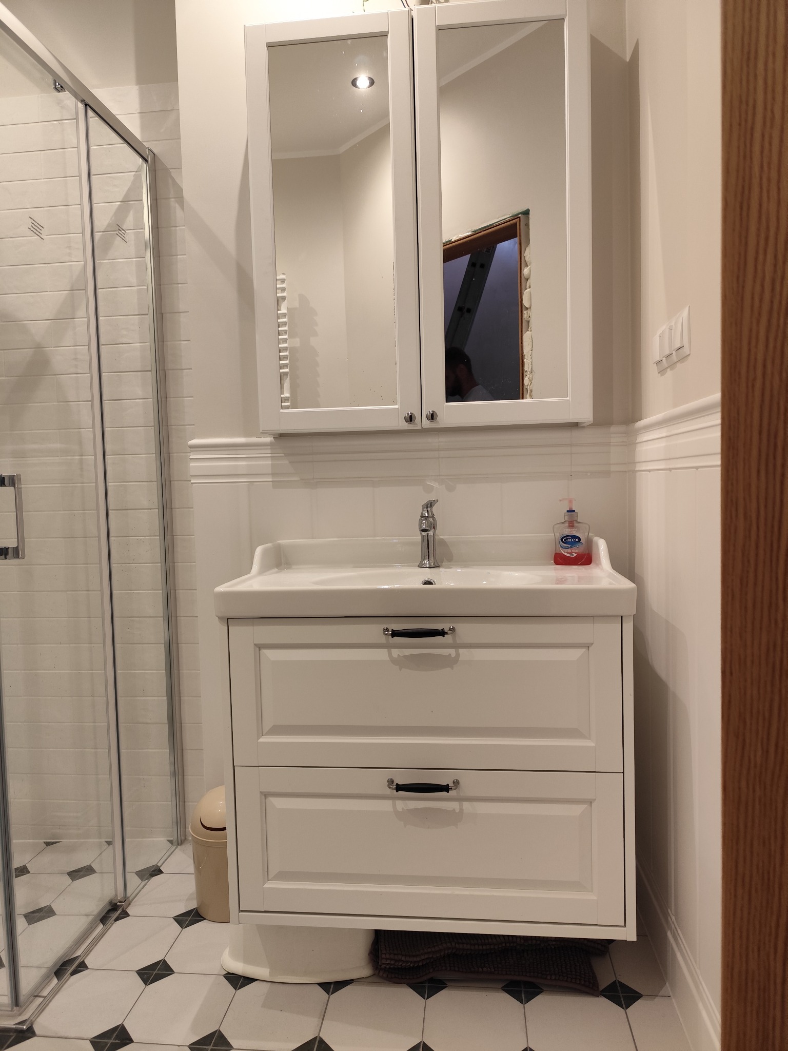 Biała szafka umywalkowa z lustrem w klasycznej łazience. Widoczna kabina prysznicowa i czarno-biała podłoga. W lustrze odbija się remontowane pomieszczenie.