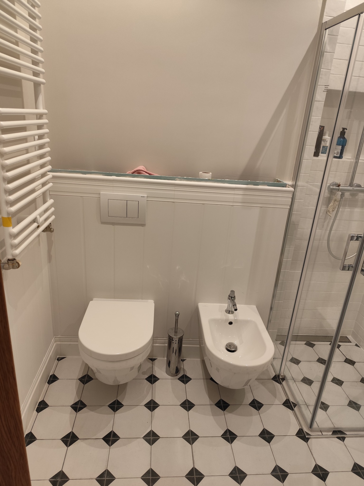 Biała łazienka z toaletą, bidetem i kabiną prysznicową. Na podłodze czarno-białe płytki w karo. Grzejnik drabinkowy po lewej, detale wykończenia ścian.