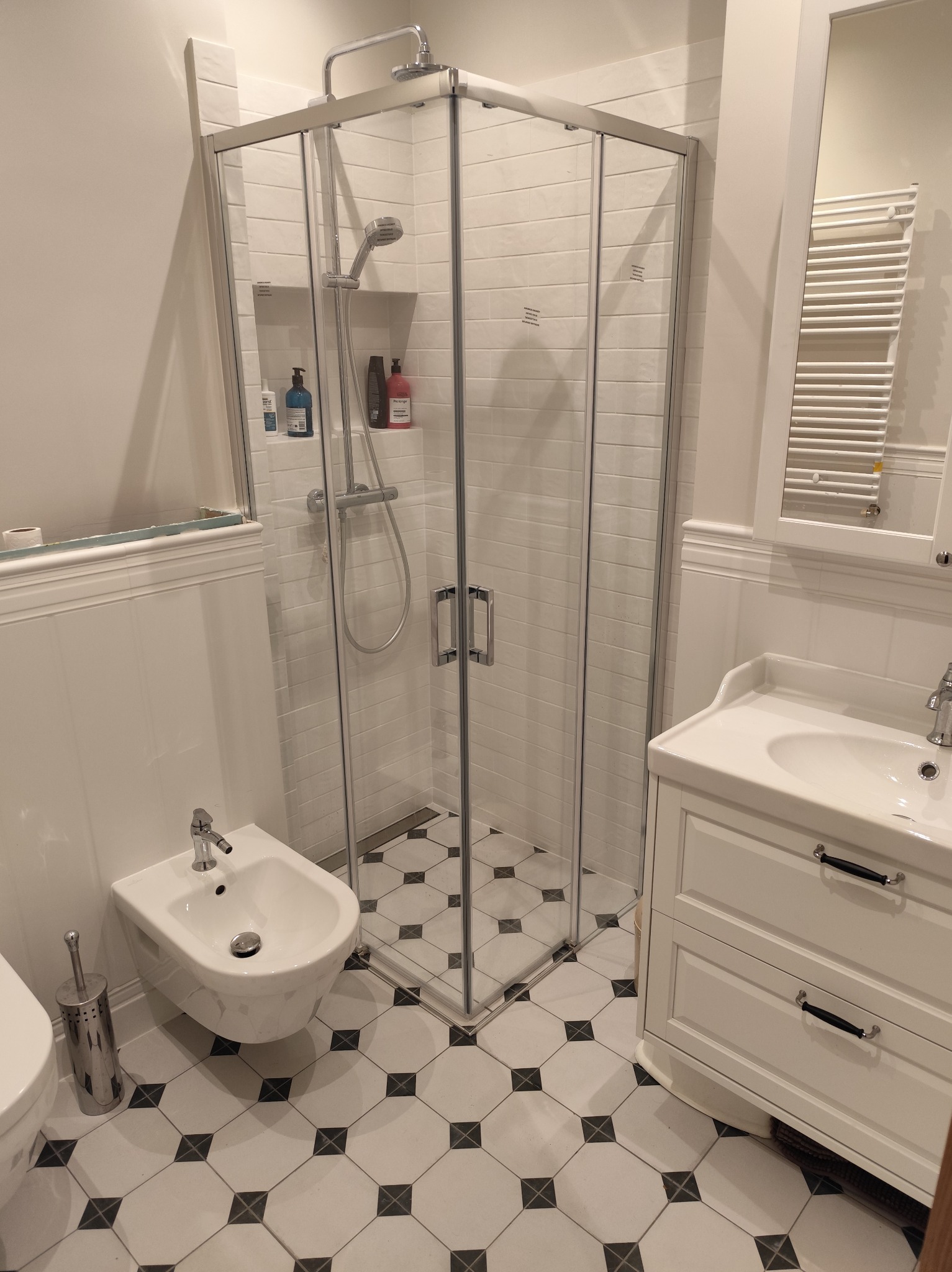 Nowoczesna łazienka z kabiną prysznicową, białą armaturą i czarno-białą podłogą w romby. Widoczny bidet, umywalka z szafką i grzejnik drabinkowy.