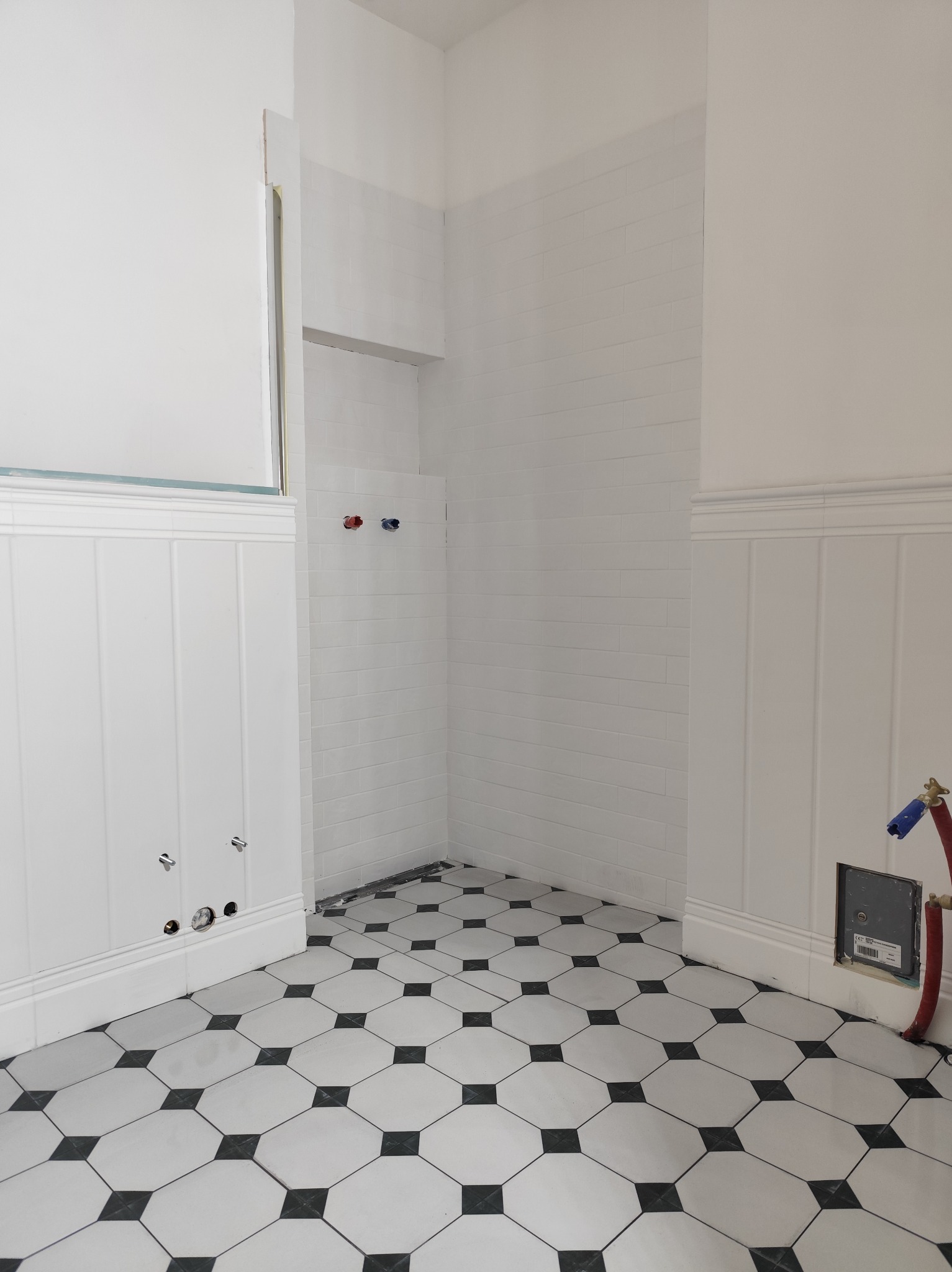 Biała łazienka z czarno-białą podłogą w karo, białe płytki na ścianach i przygotowane instalacje wodne. Widoczne pionowe panele ścienne.