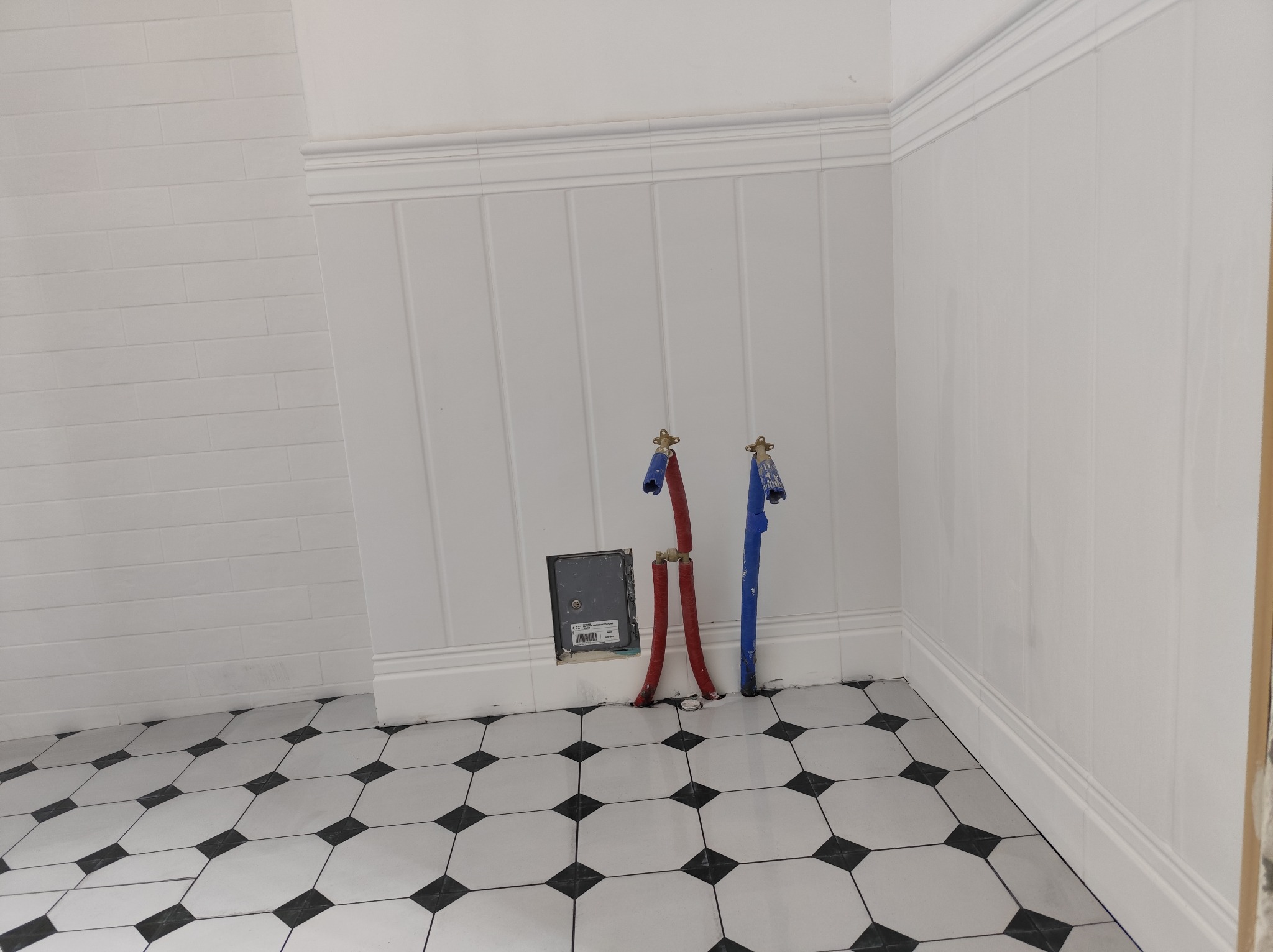 Biała łazienka z panelami ściennymi, wzorzystą podłogą i wystającymi rurami instalacji wodnej. Widoczna skrzynka rewizyjna.