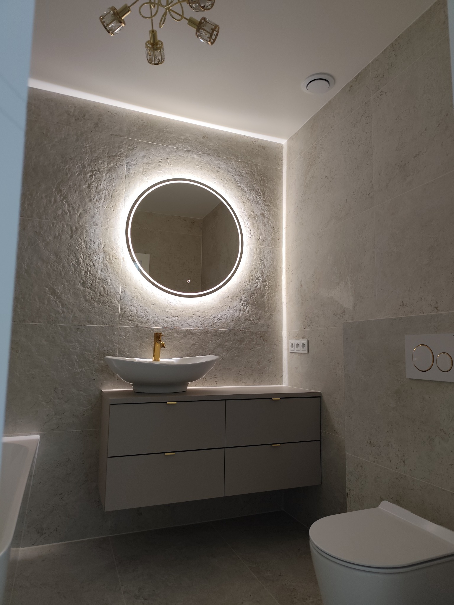 Stylowa łazienka z okrągłym lustrem LED, umywalką nablatową na szafce z szufladami i wiszącą toaletą. Płytki imitujące kamień i złote akcenty.