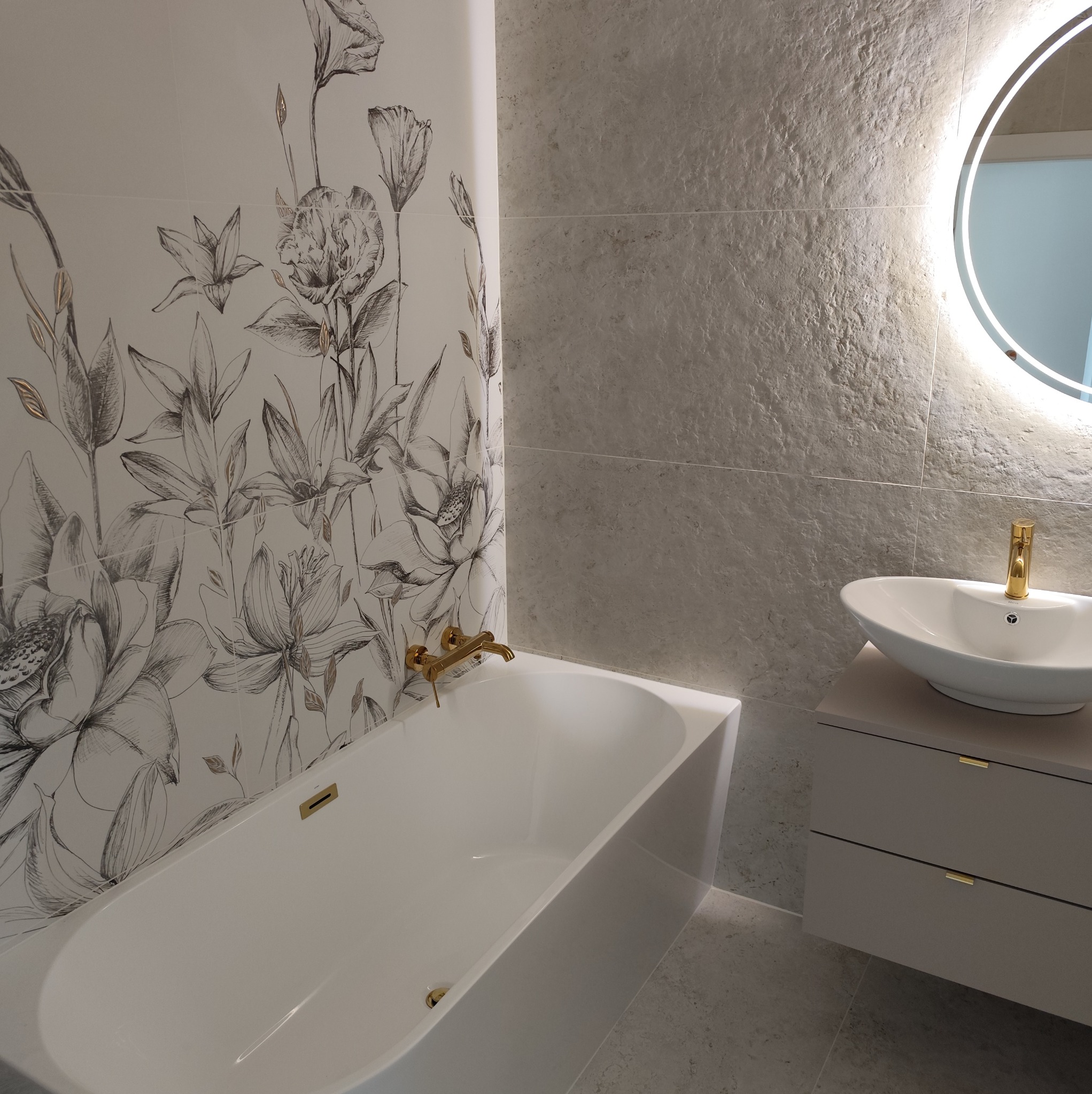 Nowoczesna łazienka z wanną, umywalką na szafce i okrągłym lustrem z podświetleniem. Ściana z płytkami w kwiatowy wzór i szare płytki imitujące kamień.