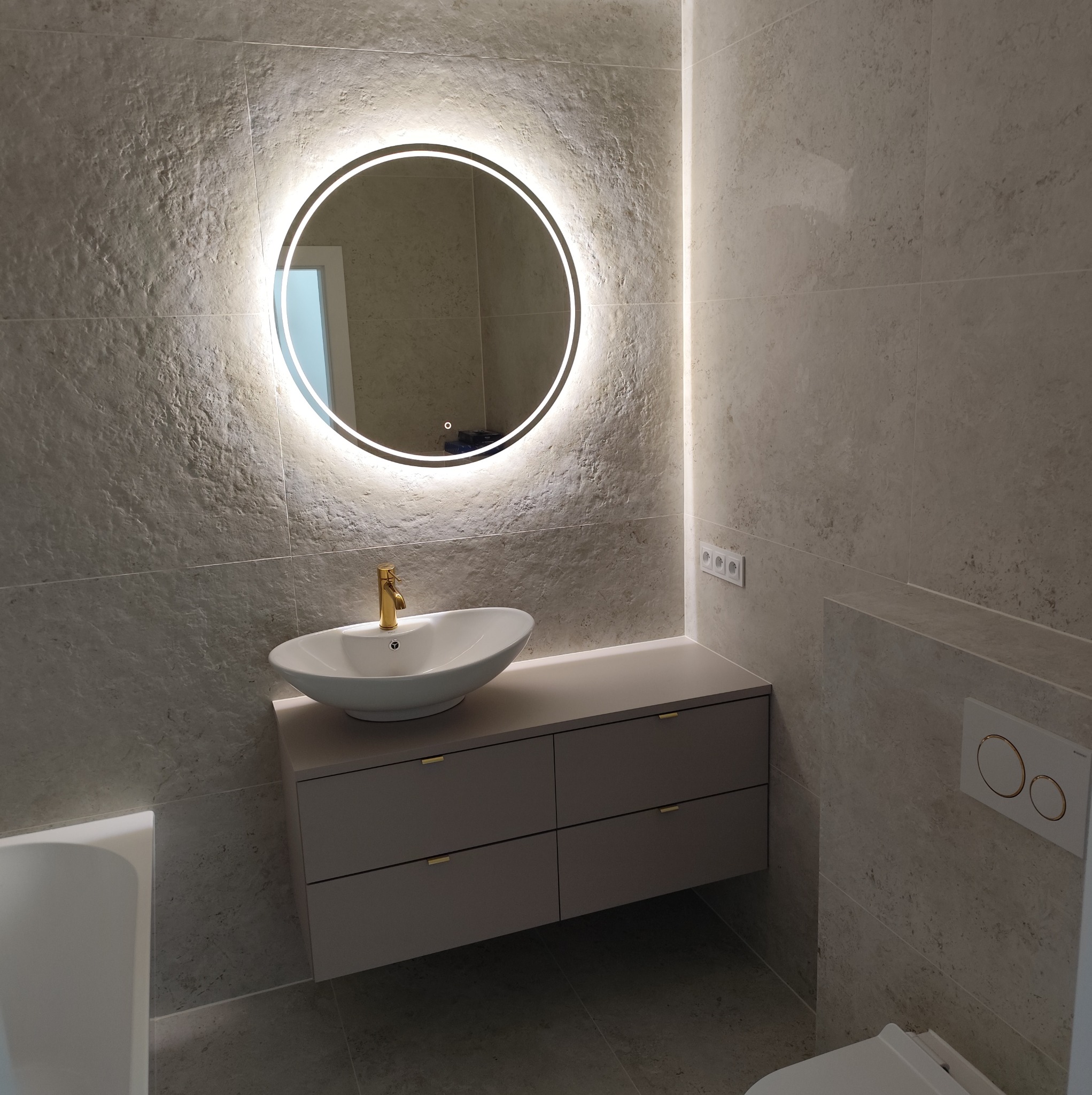 Stylowa łazienka z umywalką na szafce, okrągłym lustrem z podświetleniem LED i płytkami imitującymi kamień. Złota bateria dodaje elegancji.
