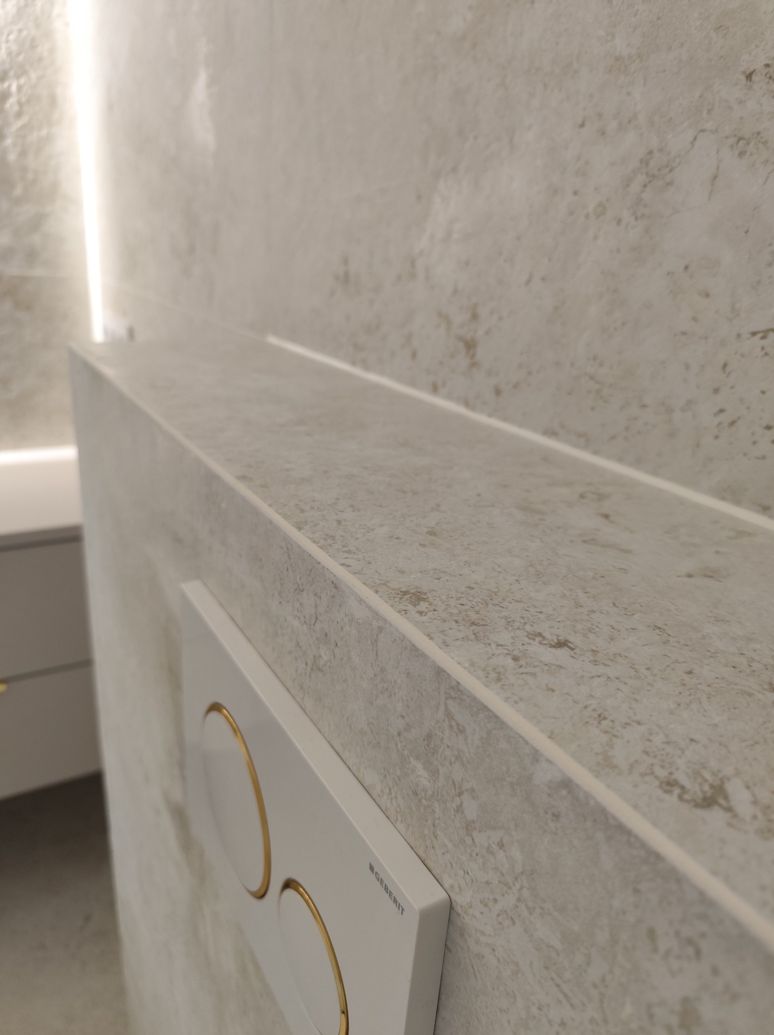 Beżowa ściana w łazience z widocznym przyciskiem spłuczki Geberit i subtelnym oświetleniem LED. Minimalistyczny design, neutralna kolorystyka.