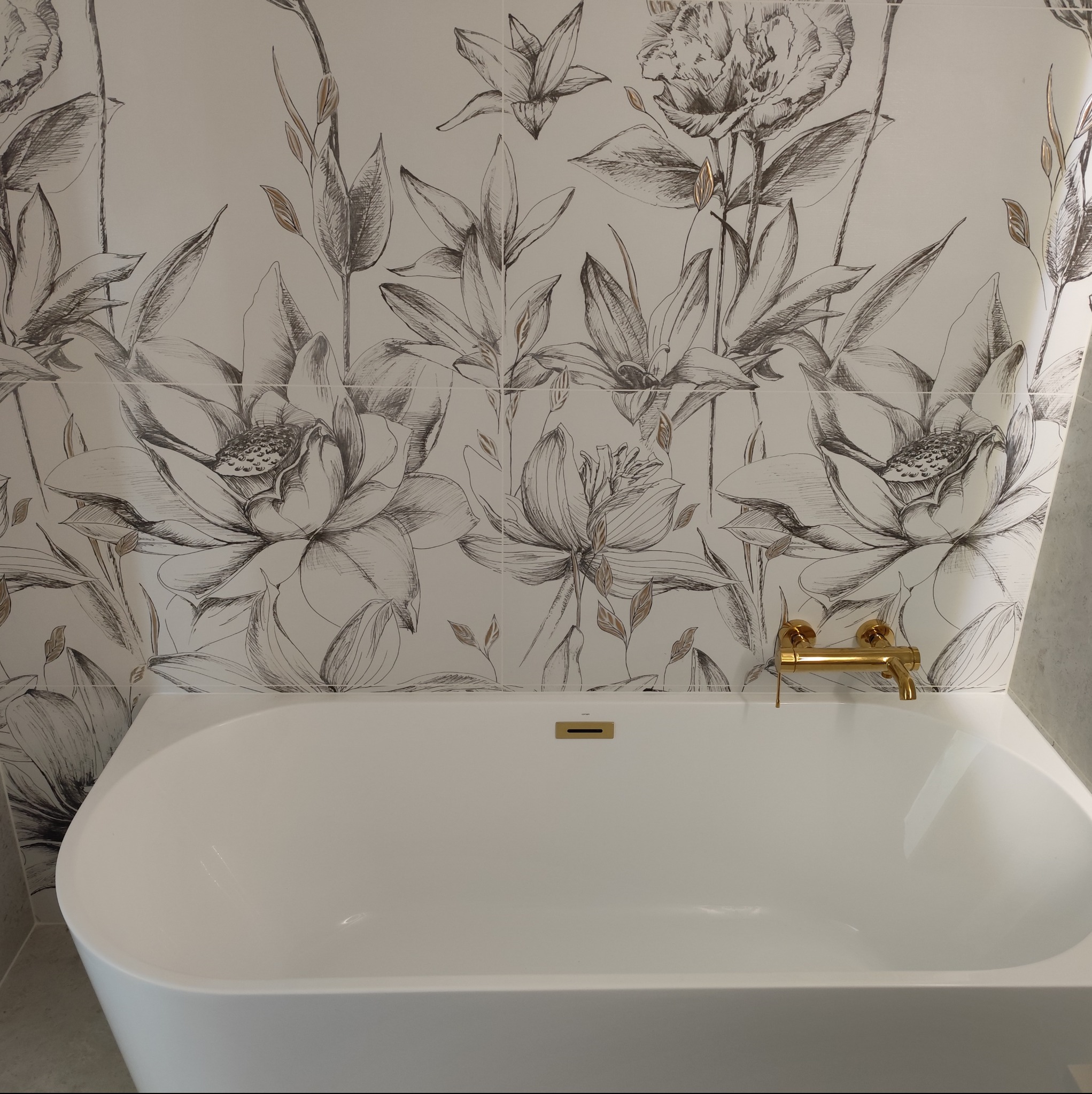 Biała wanna wolnostojąca z złotą armaturą na tle ściany z płytek w kwiatowe wzory. Nowoczesna aranżacja łazienki po remoncie.