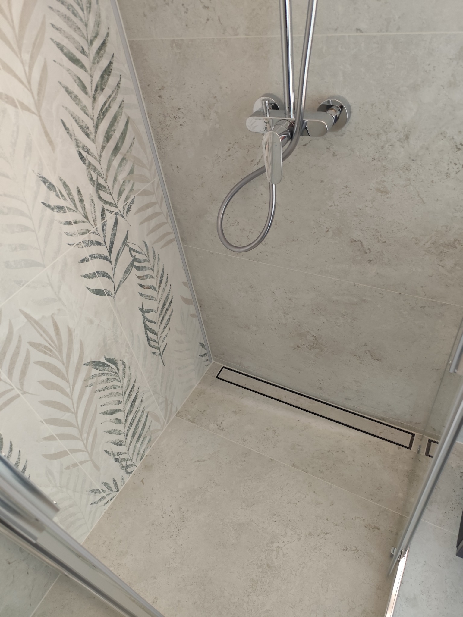 Nowoczesna kabina prysznicowa z odpływem liniowym i płytkami w odcieniach szarości, wzór liści na ścianie. Chromowana bateria natryskowa.